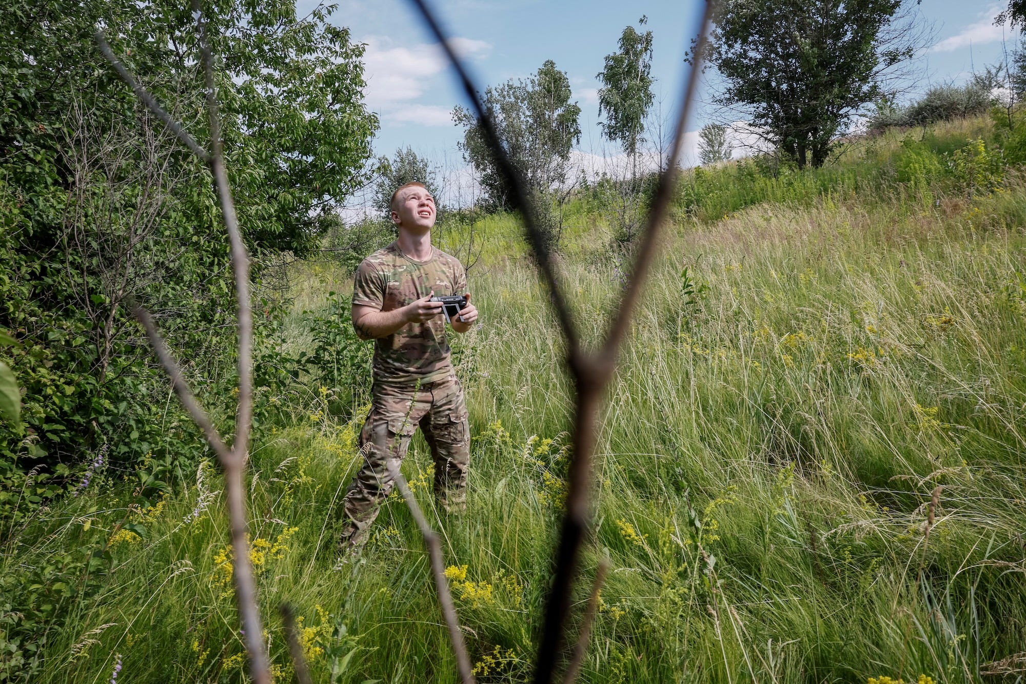 A man in a camoflage trousers and t-shirt stands in long grass holding a controller and looking up into the air