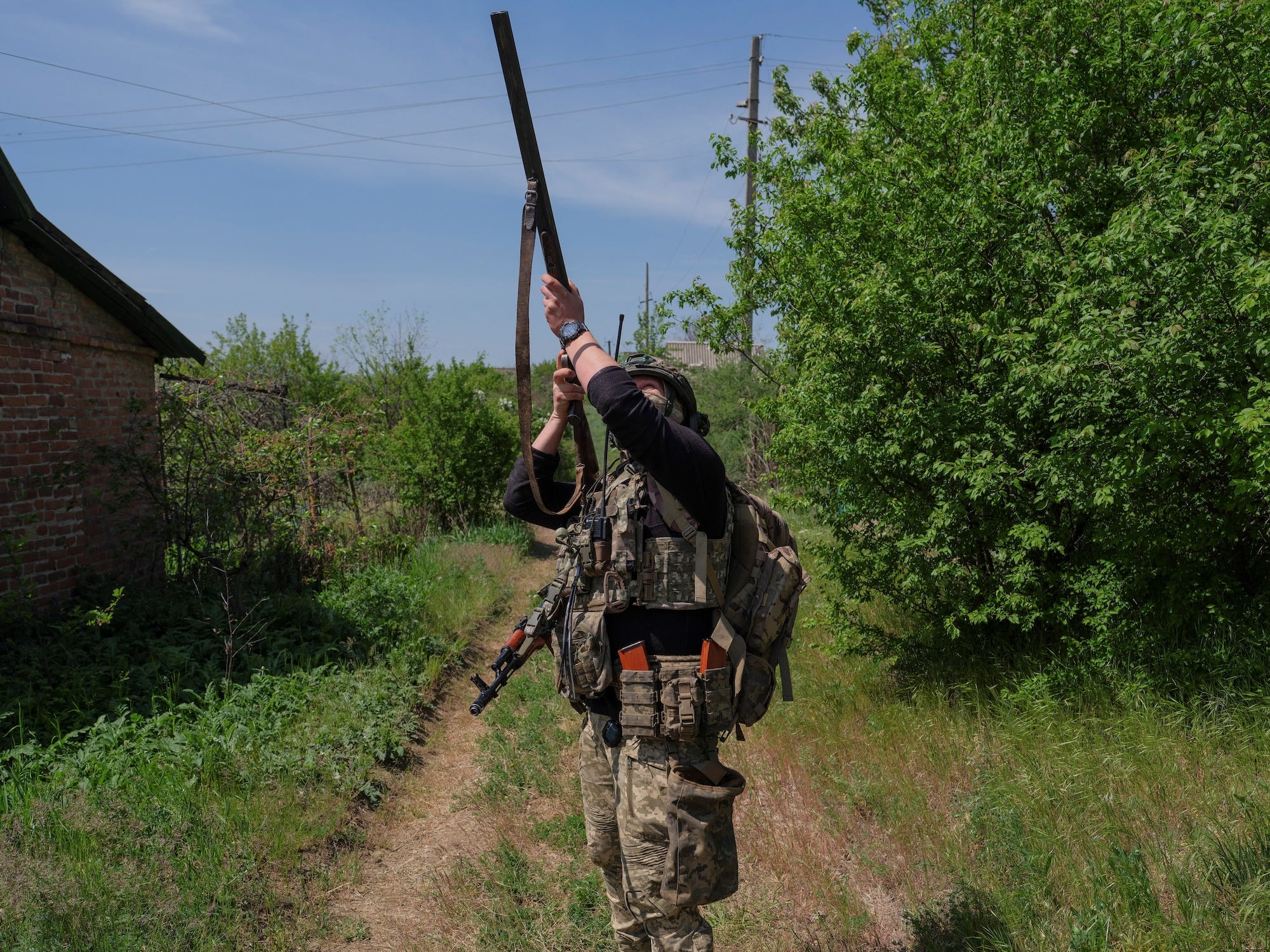 A solider wearing camoflage gear aims a shotgun into the air under a blue sky and between a brick building and a large, green bush