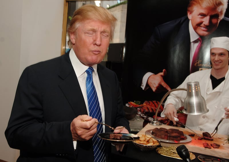 Donald Trump closes his eyes and appears to chew while holding a plate with a steak in front of a chef pointing at steak picture saying Trump Steaks