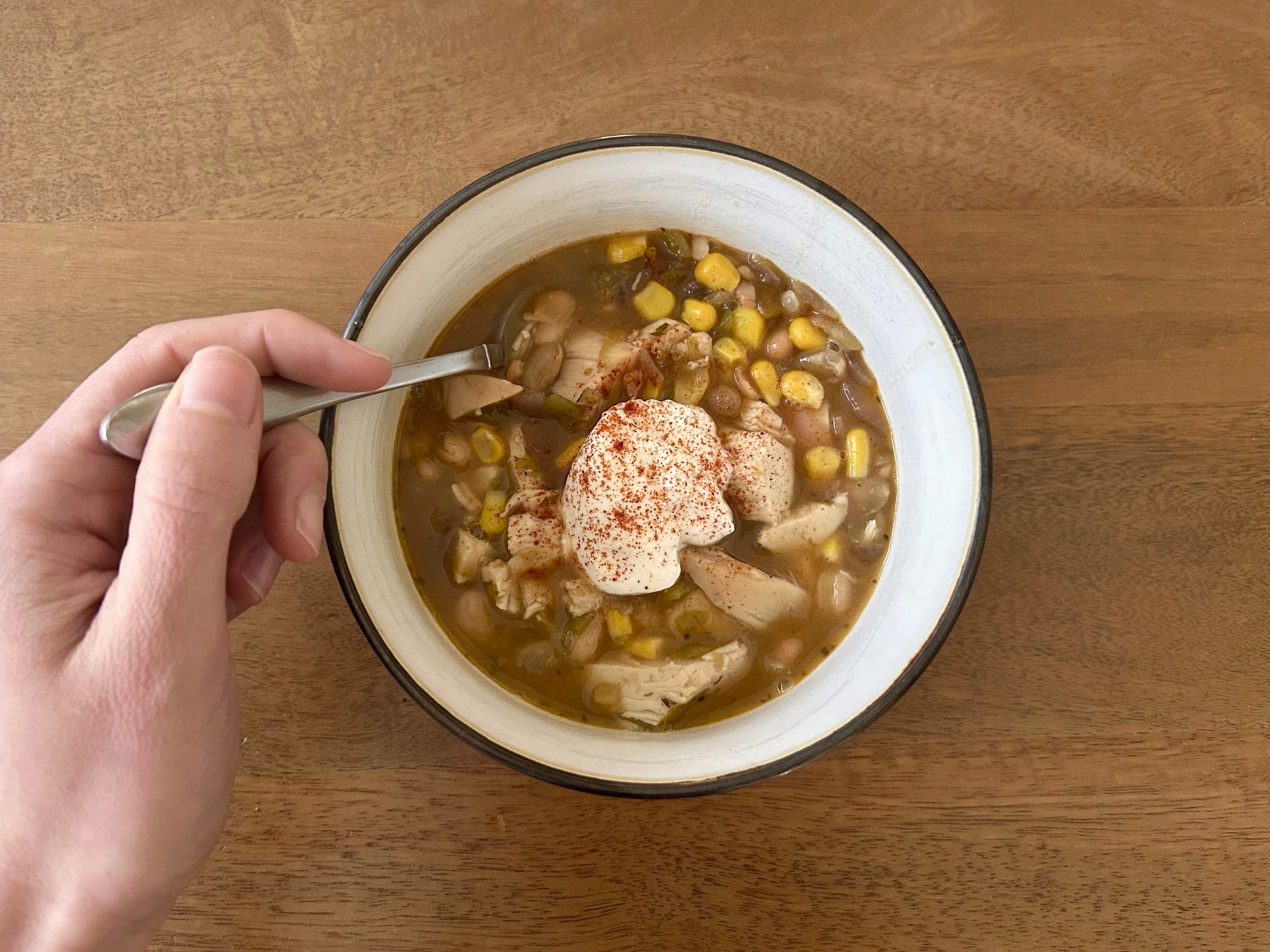 A hand holding a spoon in a bowl of chicken chili, with a dollop of sour cream on top.