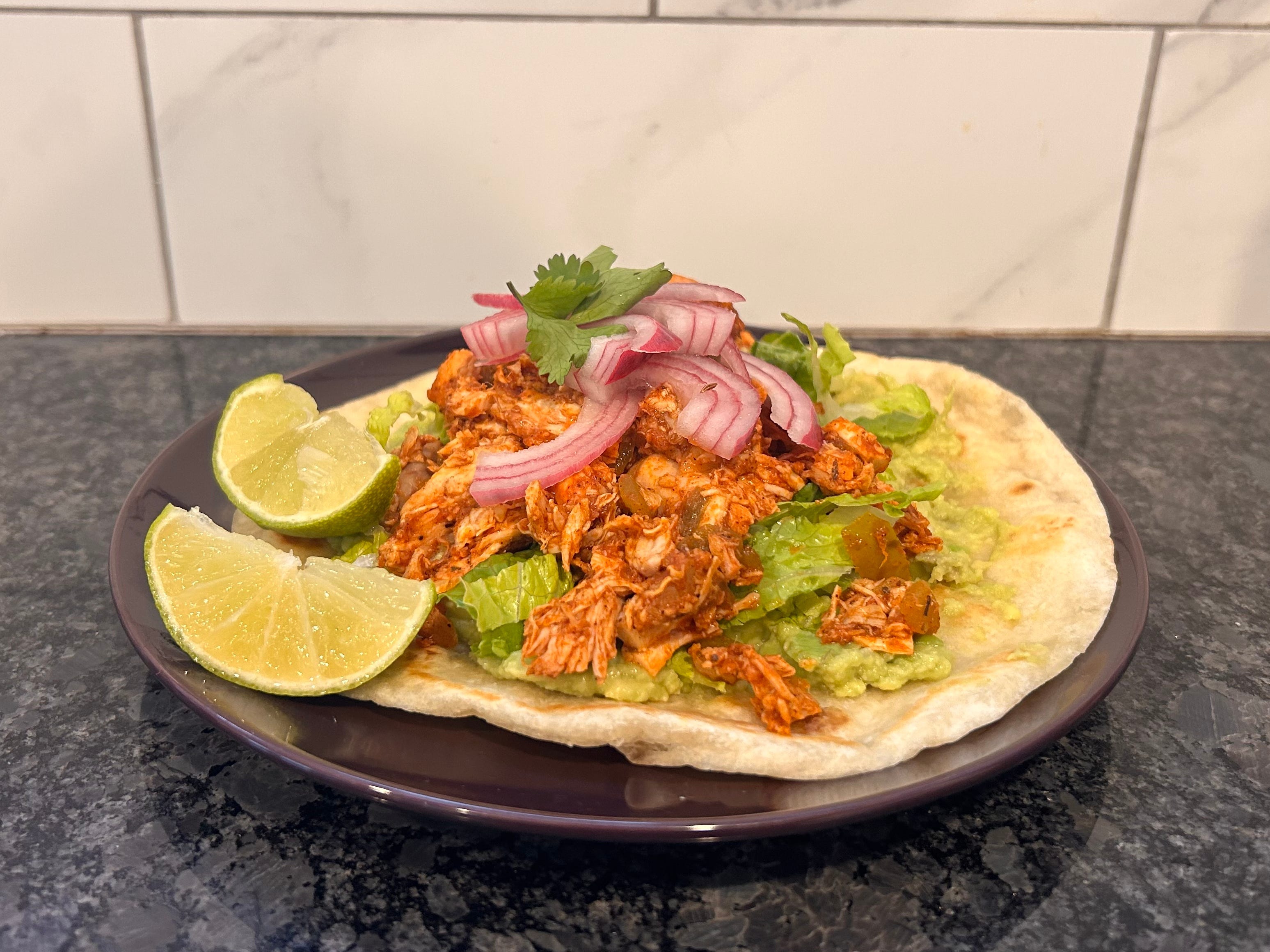 Costco rotisserie chicken on a tostada, garnished with pickled red onion and plated with two lime wedges.
