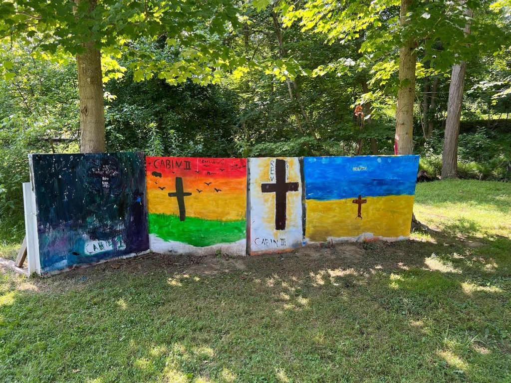 Four painted canvases depicting crosses and nature scenes stand in a grassy area.