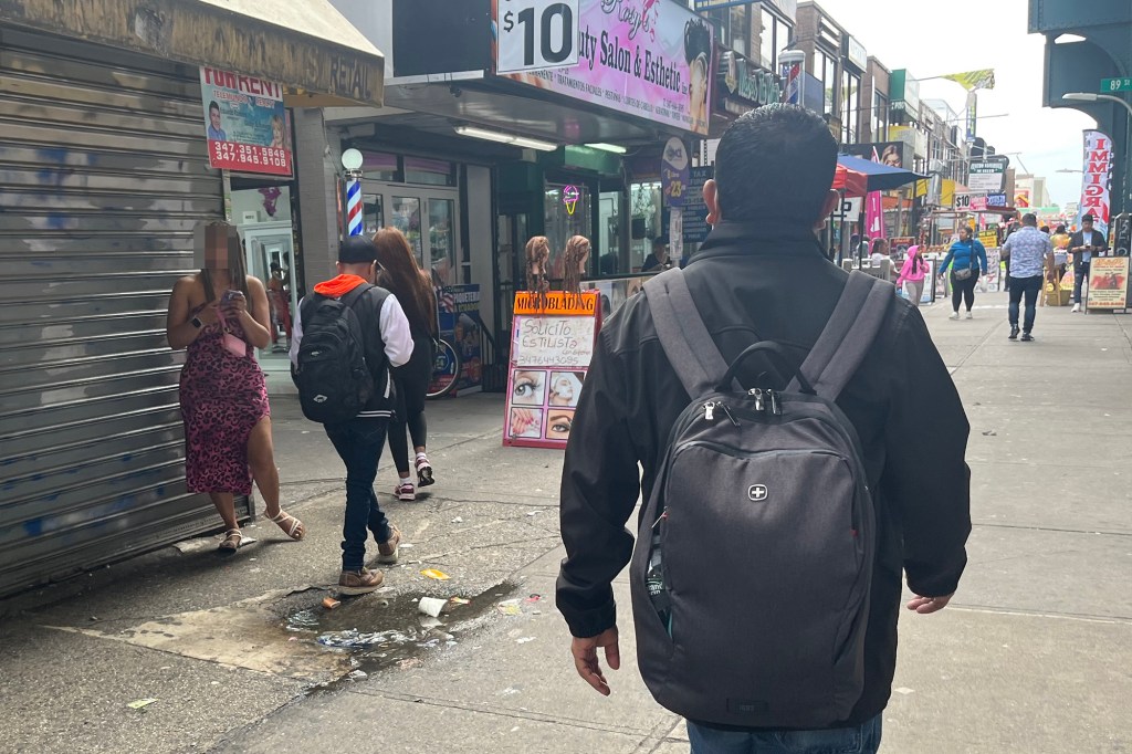 A person walks down Roosevelt Avenue in Queens, New York, past shops and pedestrians.