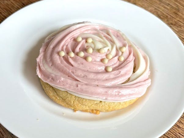 A Crumbl cookie with pink-and-white frosting and small white-pearl sprinkles on a white plate. The plate is on a wooden table.