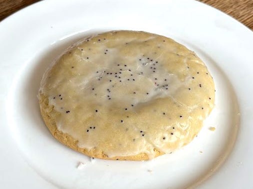 A Crumbl cookie with an icing glaze and poppy seeds on a white plate. The plate is on a wooden table.