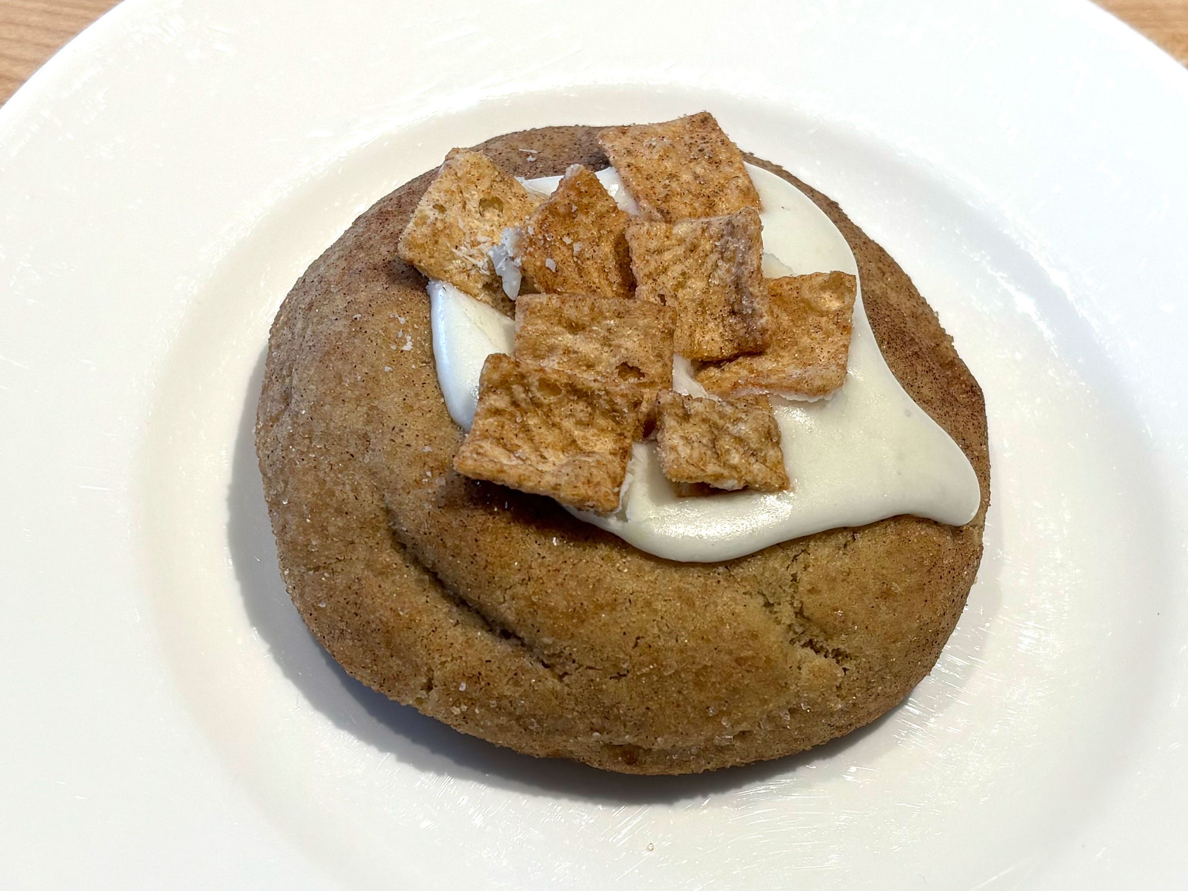 A cookie topped with cream-cheese icing and pieces of cinnamon cereal on a white plate.