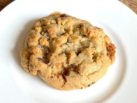 A Crumbl cookie with milk-chocolate chips and toffee on a white plate. The plate is on a wooden table.