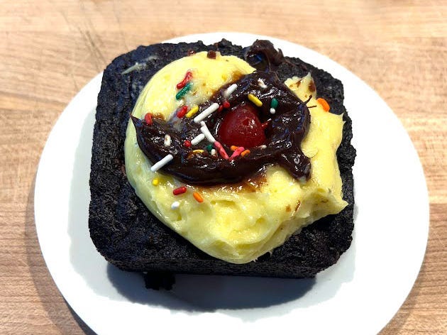 A brownie topped with yellow frosting, chocolate fudge, rainbow sprinkles, and a cherry on a white plate.