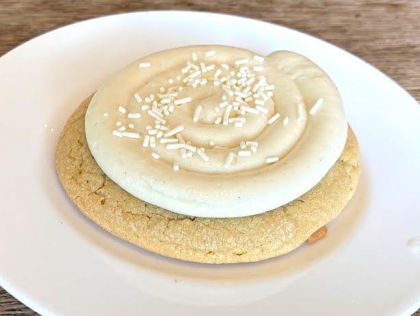 A vanilla cookie topped with white icing and white sprinkles on a white plate.