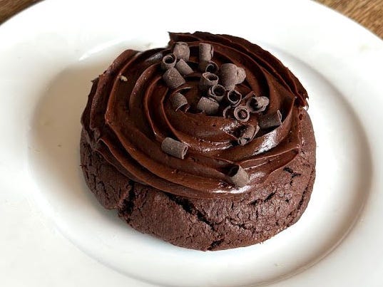 A chocolate Crumbl cookie with chocolate frosting and rolled-chocolate shavings on a white plate. The plate is on a wooden table.