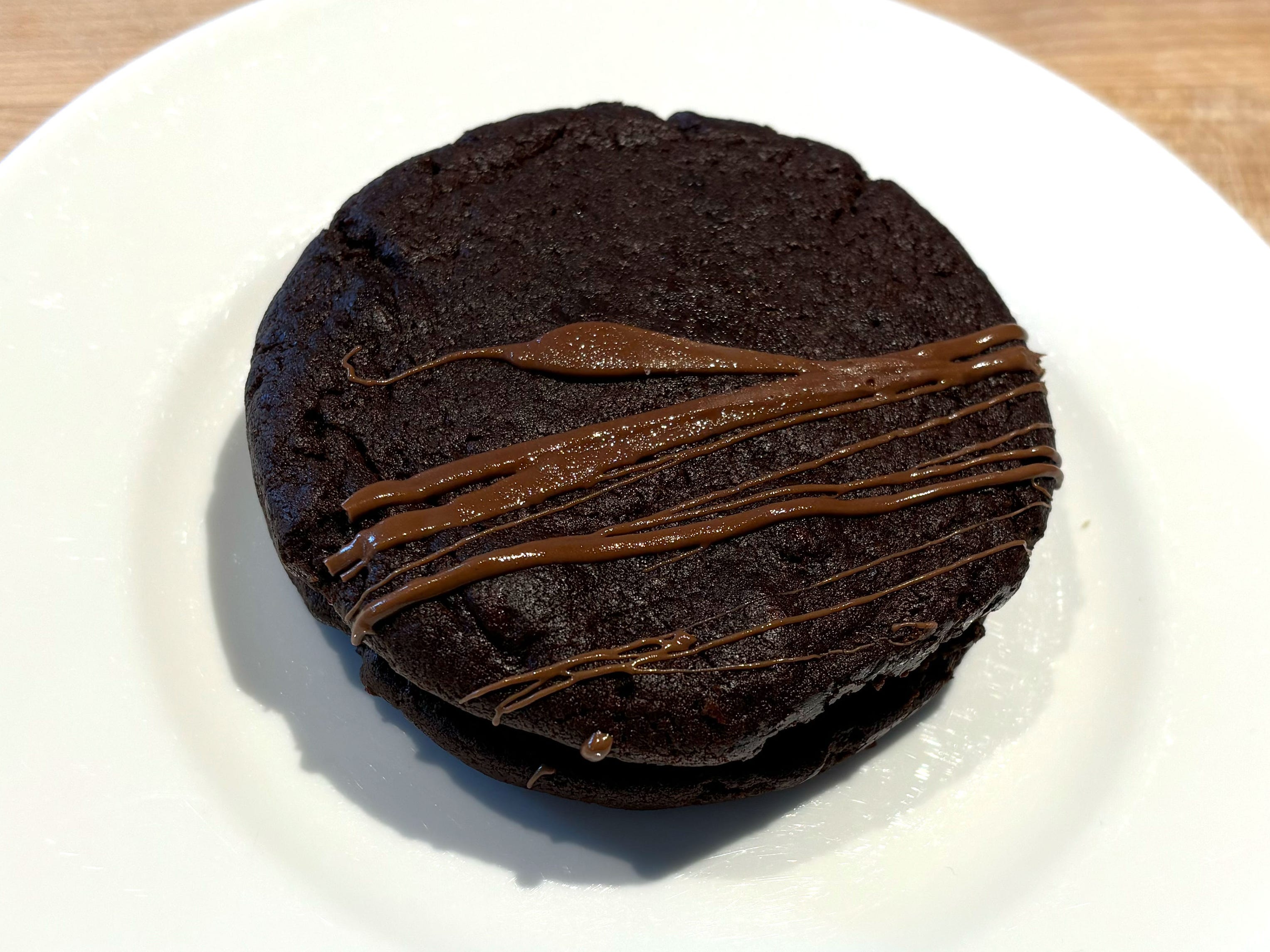 Two chocolate cookies with fudge in the middle and chocolate drizzled on top. The cookie is on a white plate.
