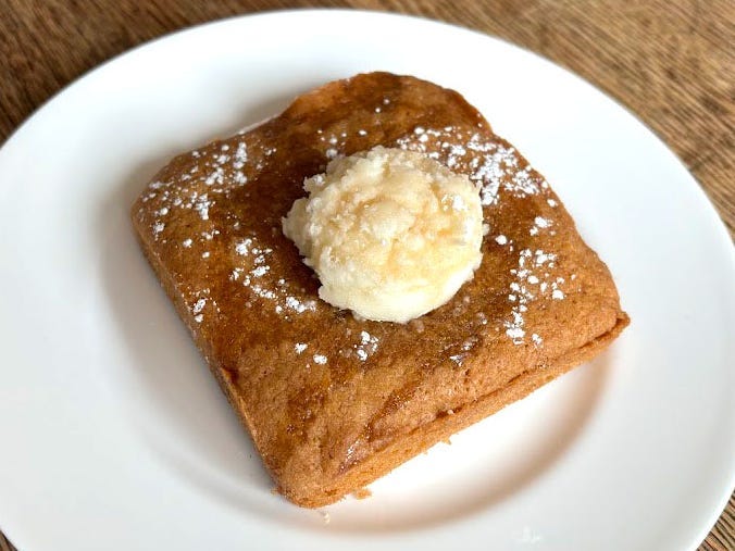 A piece of rectangular Crumbl cheesecake with a dollop of icing and powdered sugar on top. The cheesecake is on a white plate placed on a wooden table.