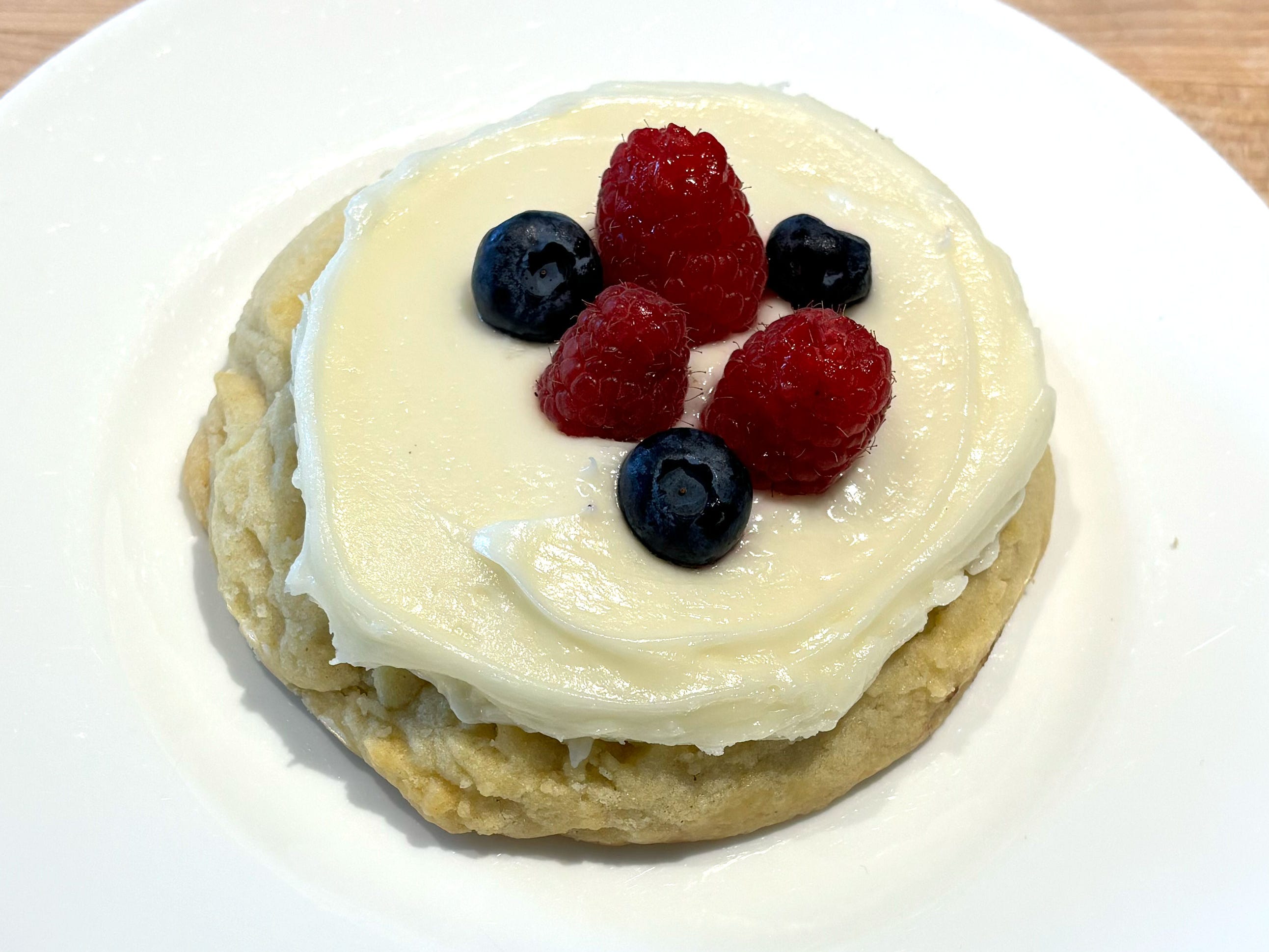 A vanilla cookie topped with vanilla icing, raspberries, and blueberries. The cookie is on a white plate.