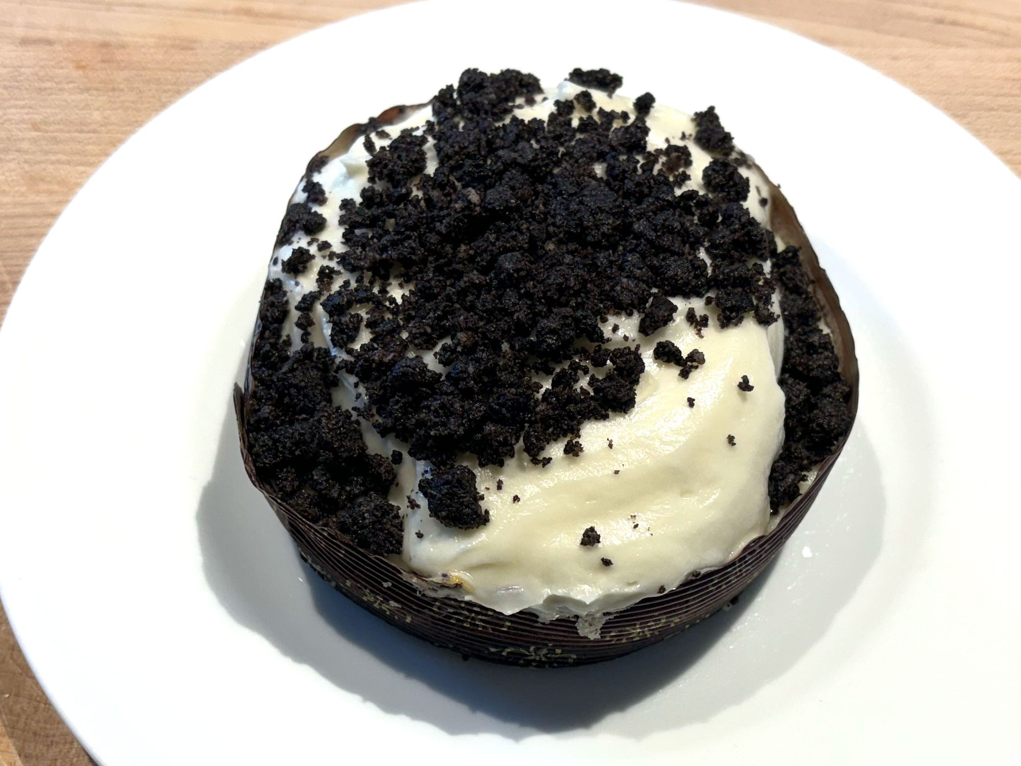 A circular piece of cookies-and-cream cheesecake topped with whipped cream and cookie pieces. The cheesecake is on a white plate.
