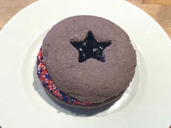 A purple cookie sandwich with a star cut-out on top that's filled with jelly. Between the two cookies are icing and red, white, and blue sprinkles.