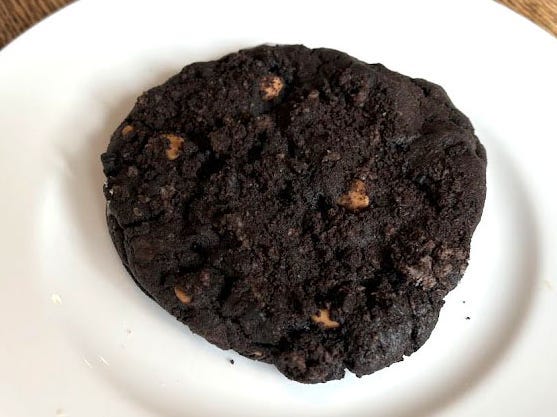 A chocolate Crumbl cookie with peanut-butter chips on a white plate. The plate is on a wooden table.