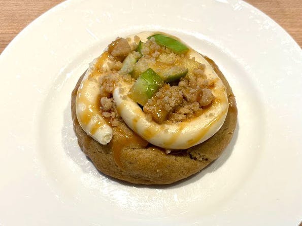 A cookie topped with a swirl of white icing, crumbs, and pieces of green apple on a white plate.