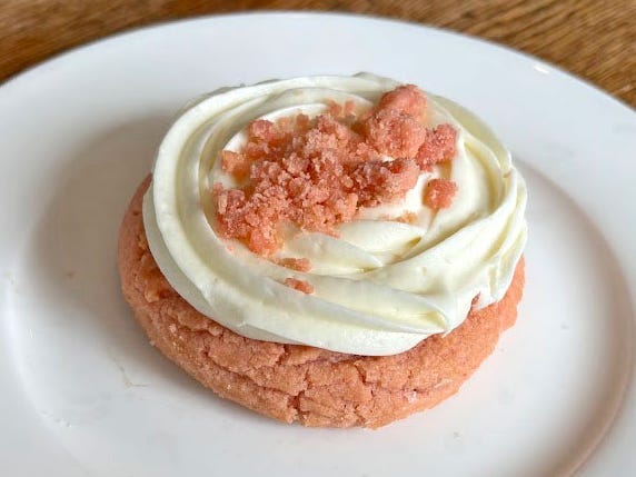 A pink Crumbl cookie with white frosting and pink crumbles on top. The cookie is on a white plate on a wooden table.