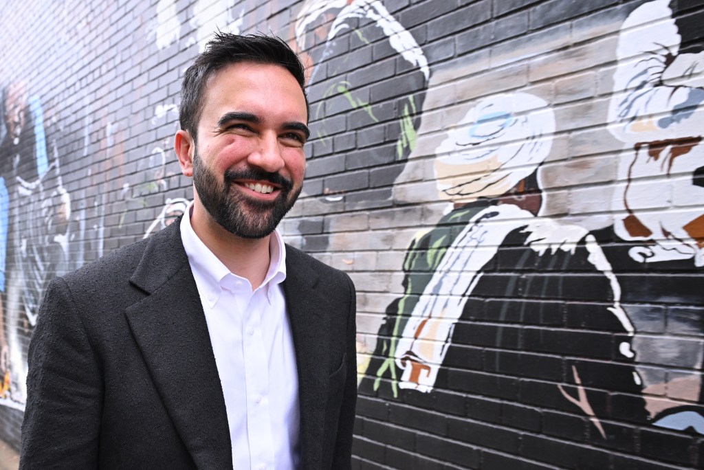 Zohran Mamdani, NYC mayoral candidate, smiling in front of a brick wall with a mural.
