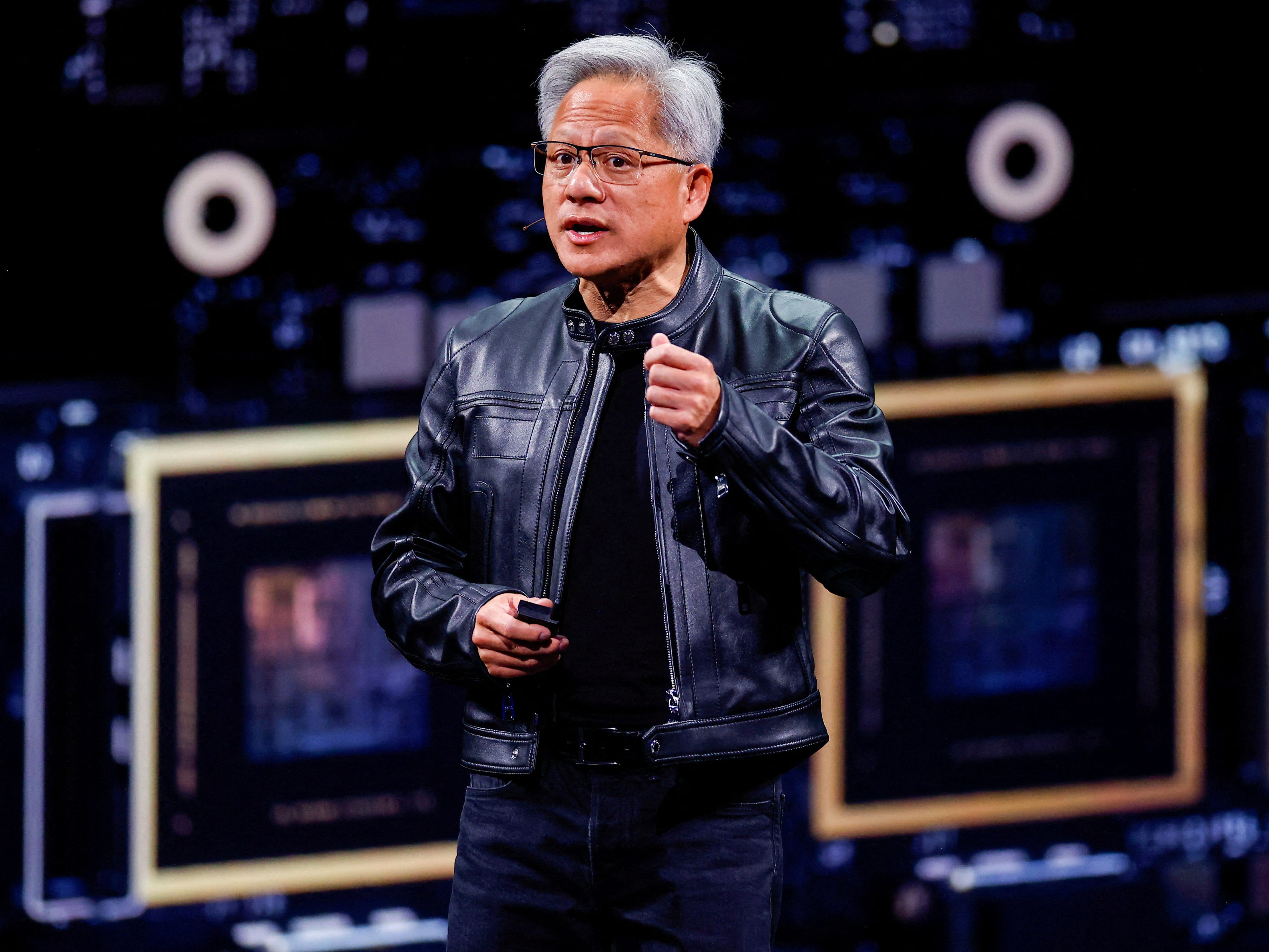 A man (Jensen Huang) wearing all black and a black leather jacket speaks on a stage in front of an enlarged image of a graphics processing unit.
