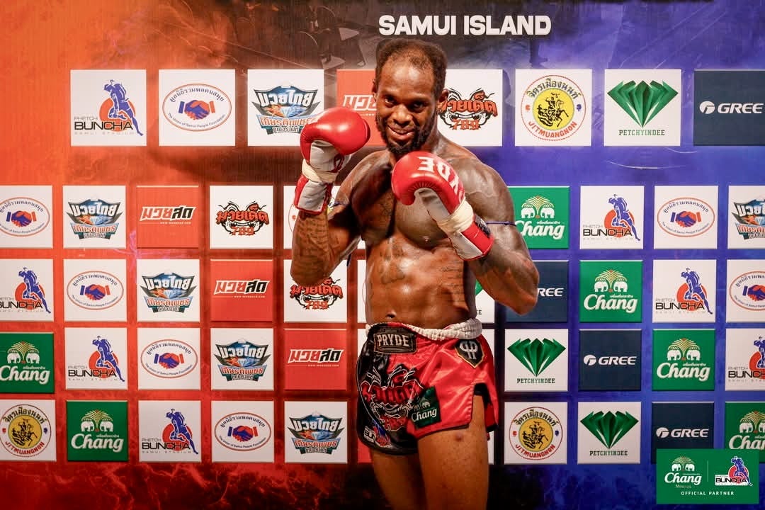 A man posing in boxing gloves in front of a colorful sign after fighting in a Muay Thai match in Koh Samui, Thailand.