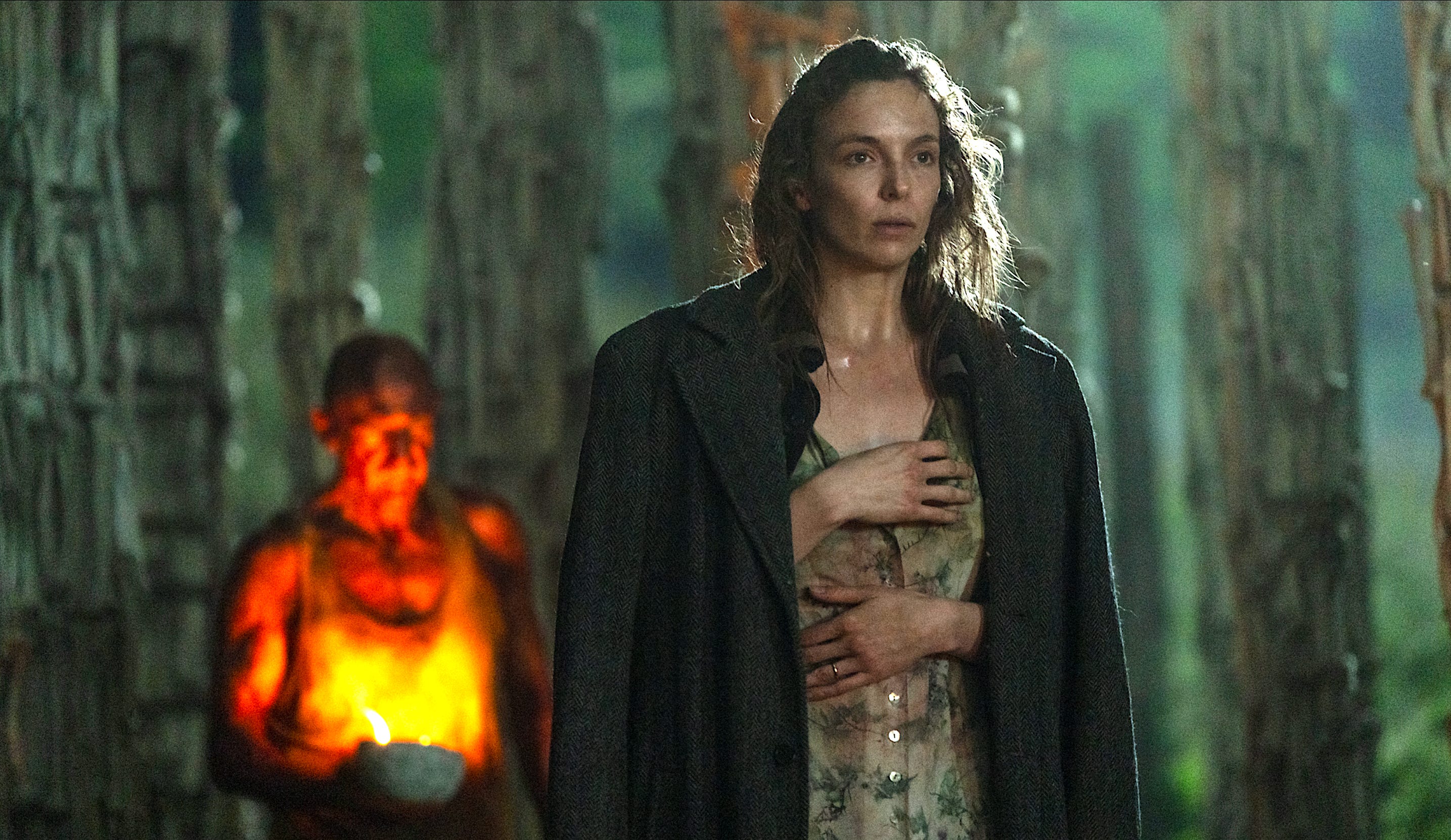 A man and a woman stood in a forest while surrounded by large poles made out of bones. On the left, a bald man is wearing a white vest and is illuminated by a candle that he's holding, but he is out of focus. On the right, the woman has messy brown hair and is wearing a large gray coat over the top of a stained floral dress. She has her hands over her chest and ribs.