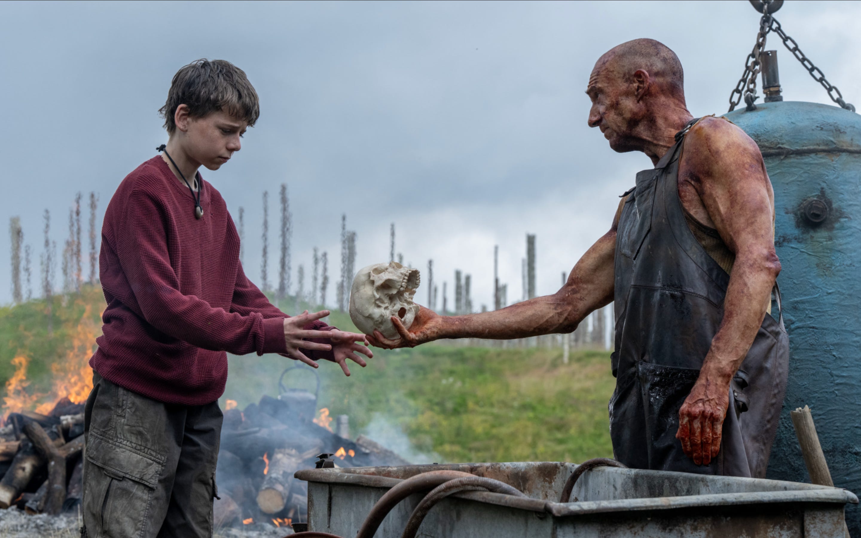 An older bald man hands a teenage boy a skull. On the left, the teenager is wearing a red sweater with a string necklace and plaid cargo pants. On the right, the older man is wearing a black apron and is handing the skull over to the boy with his right hand. Behind them, there is a pile of burning logs.