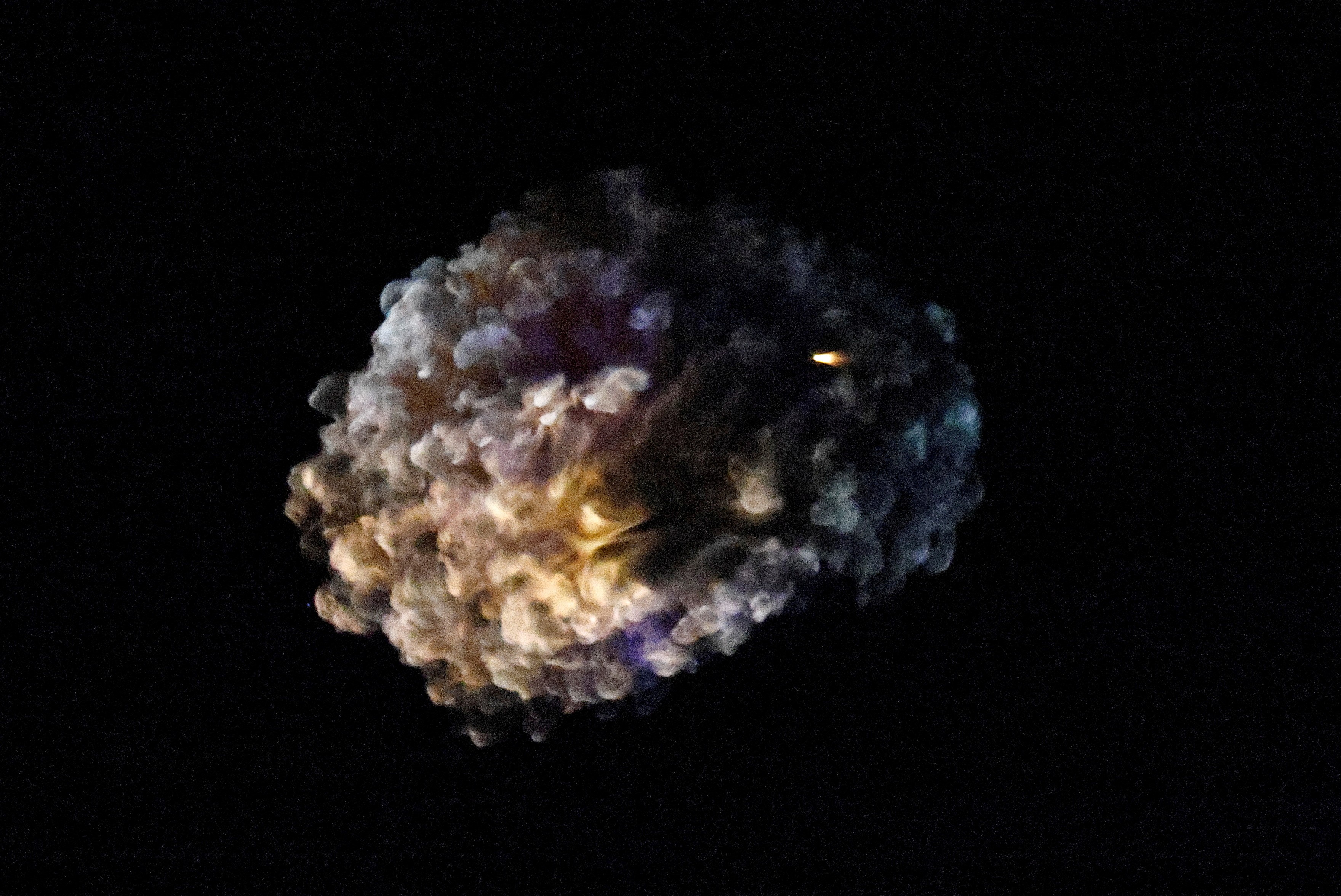 A multicolored cloud, left by an explosion, hangs in the night sky.
