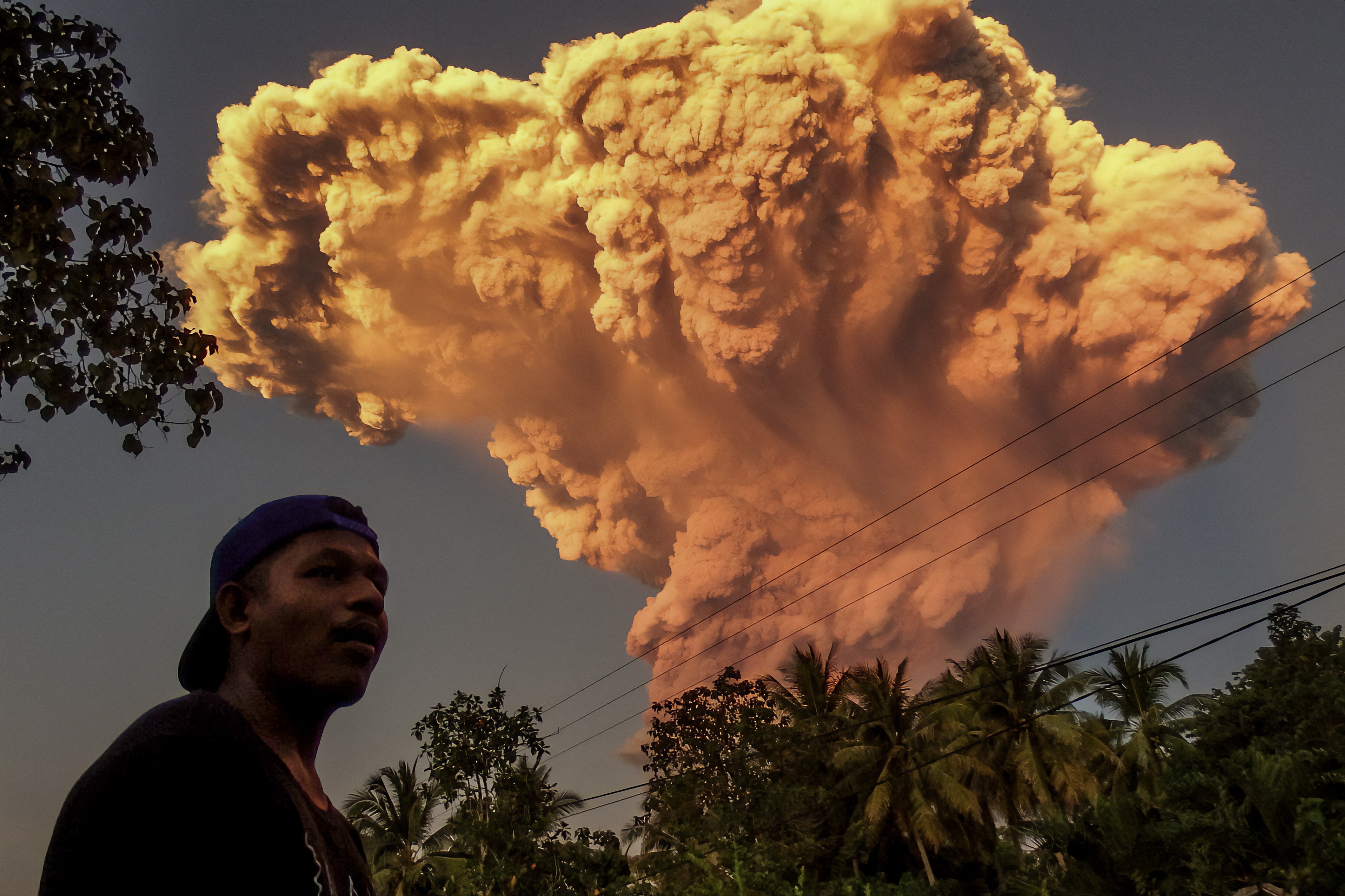 A person stands on a street, while behind them a tall ash cloud rises into the sky from a volcano.