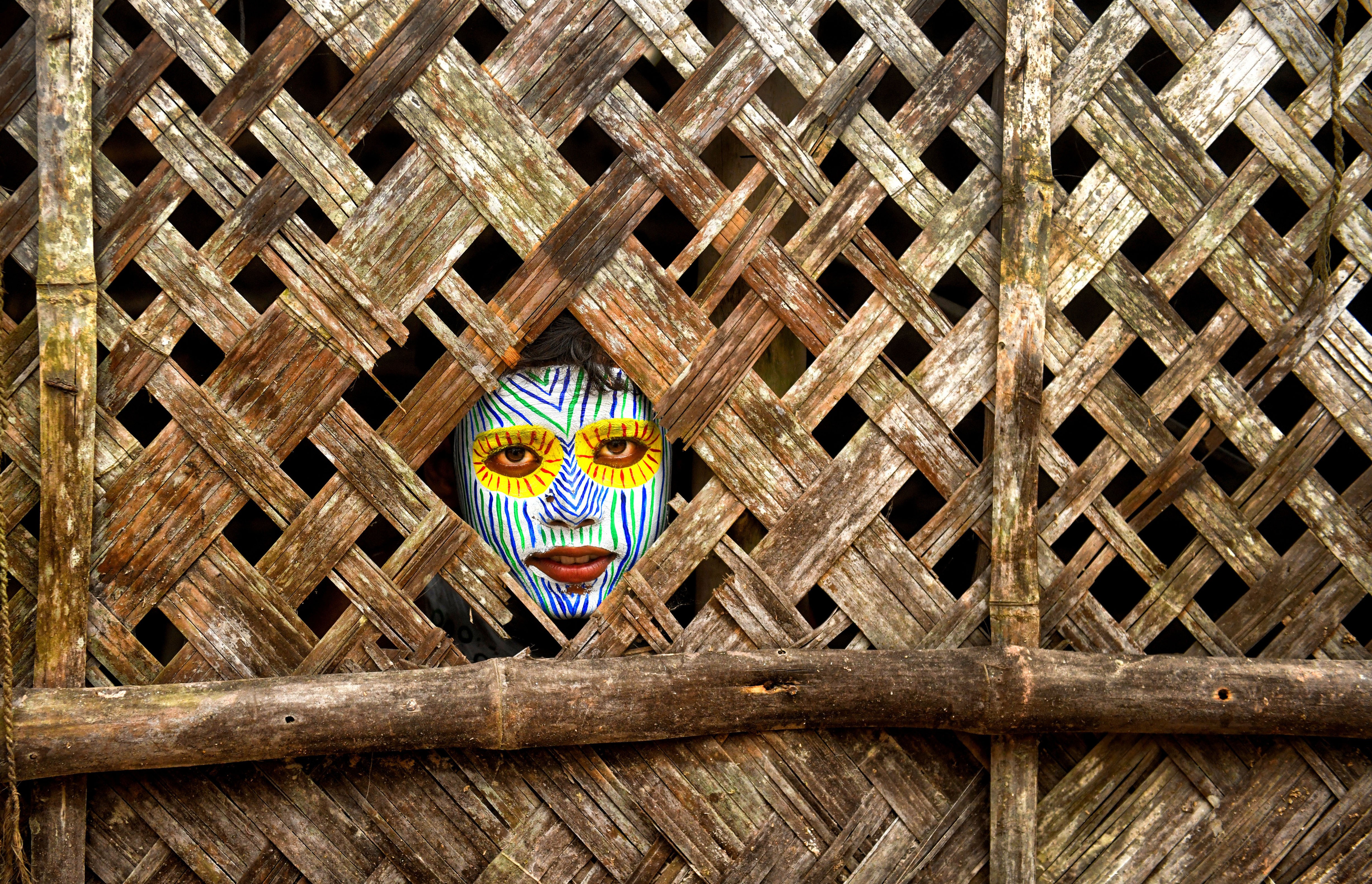 A child with their face painted peeks through a hole in a wall made of woven boards.