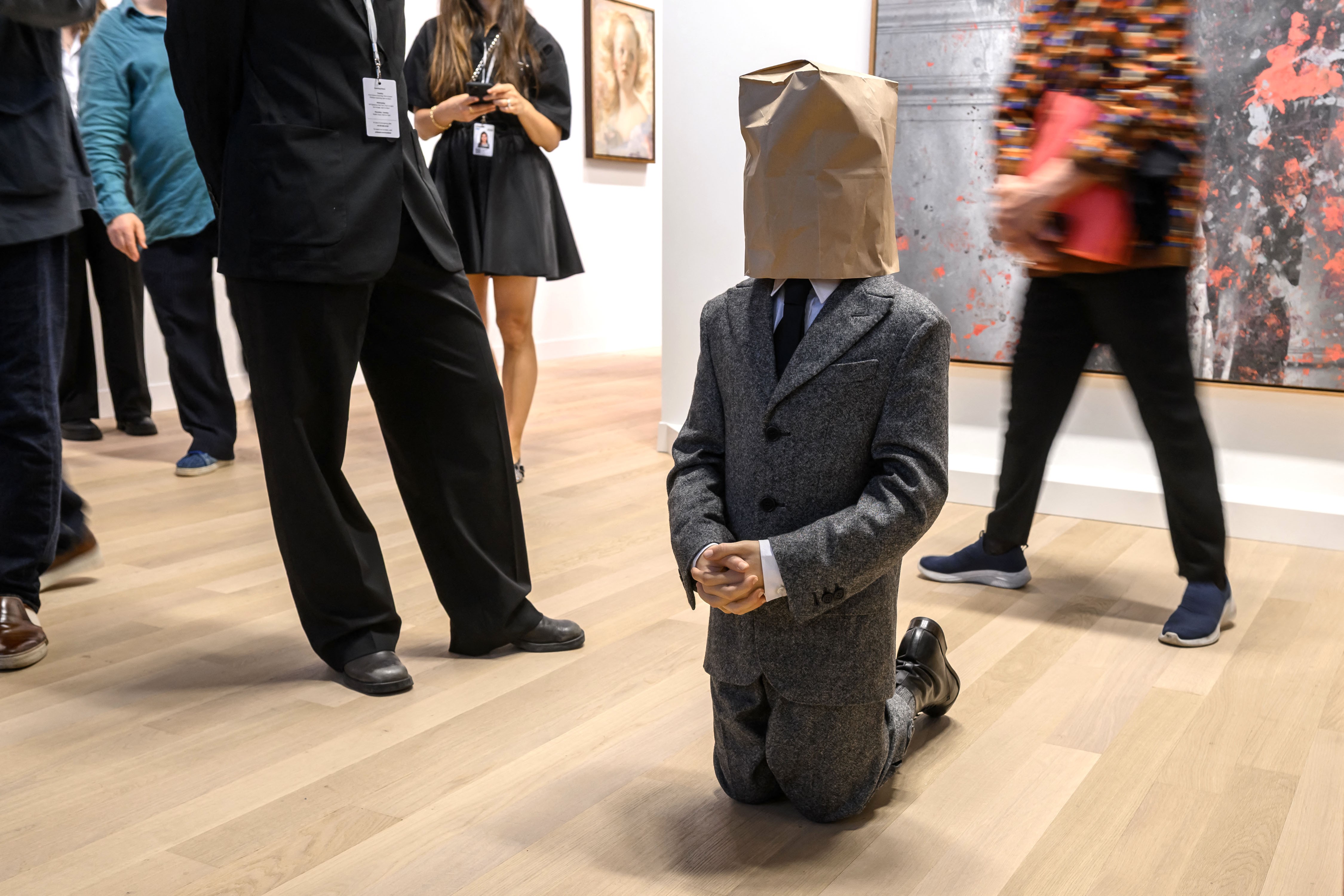 In an art gallery, people walk about and look at artwork, including a small-scale statue of a man wearing a suit that is depicted as kneeling while wearing a paper bag over its head.
