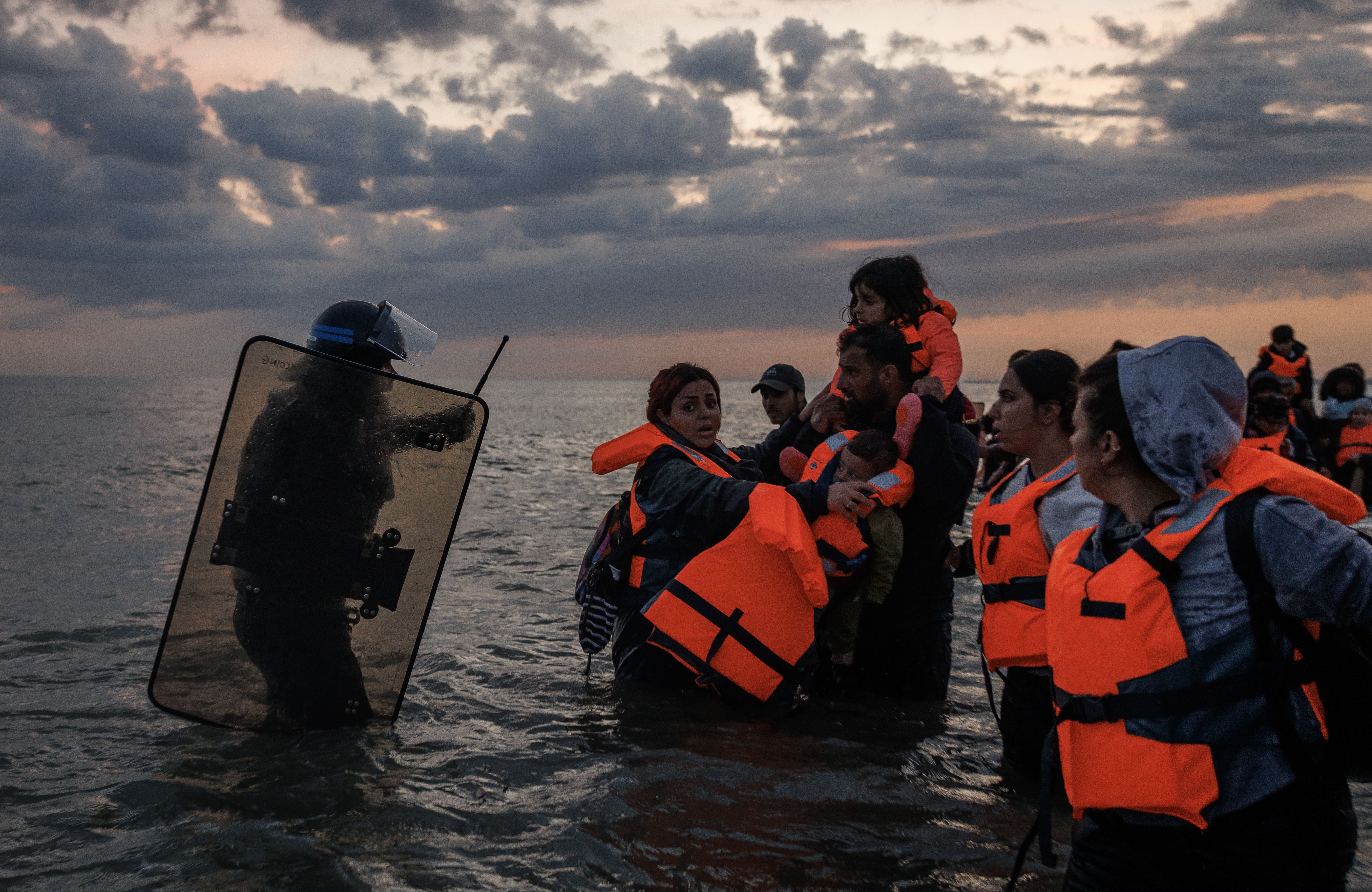 A riot police officer carrying a shield and baton walks into knee-deep water toward a group of men, women, and children who are wearing life jackets.