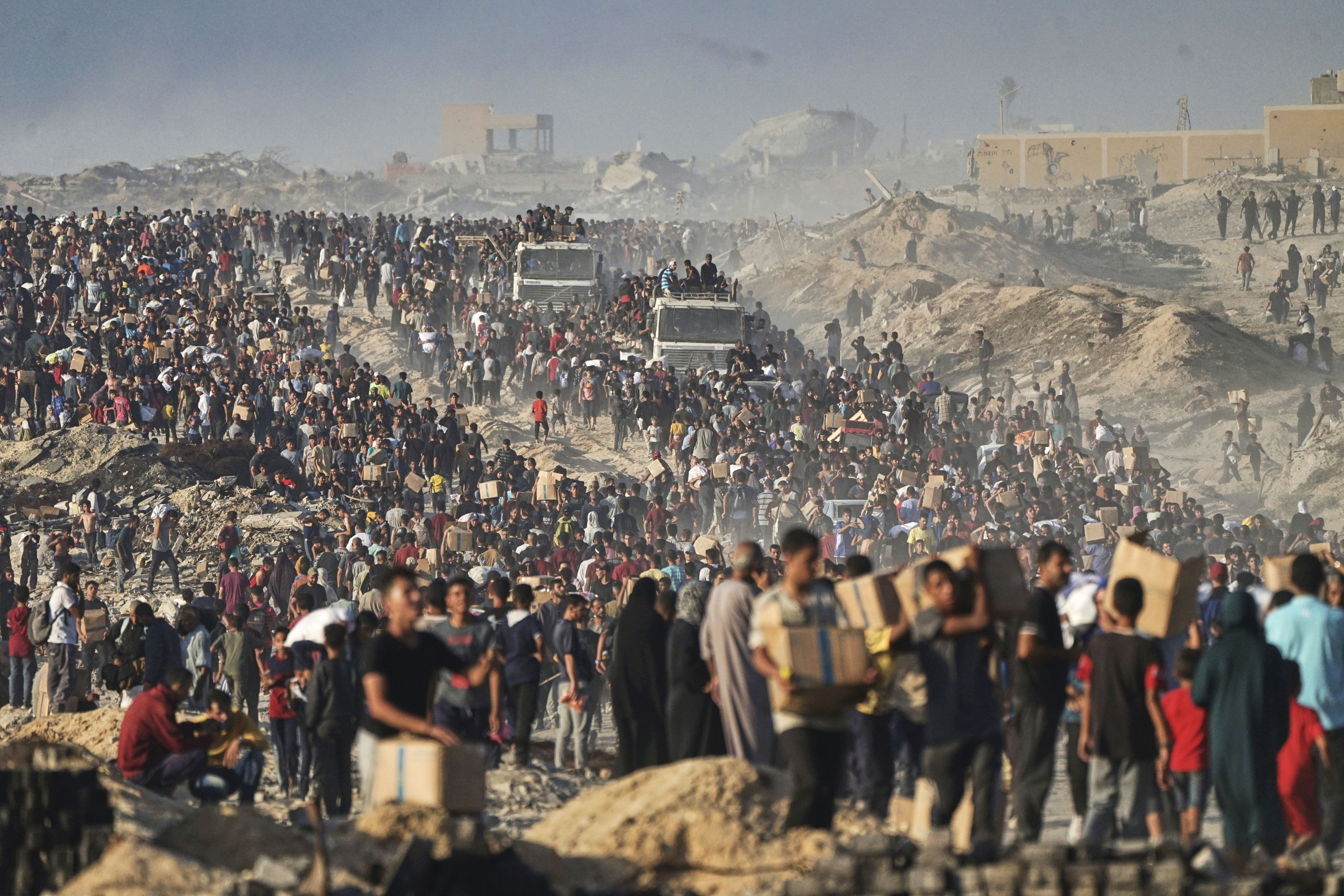 A large crowd of Palestinians walks along a dirt road, many of them carrying boxes and sacks of food and supplies.