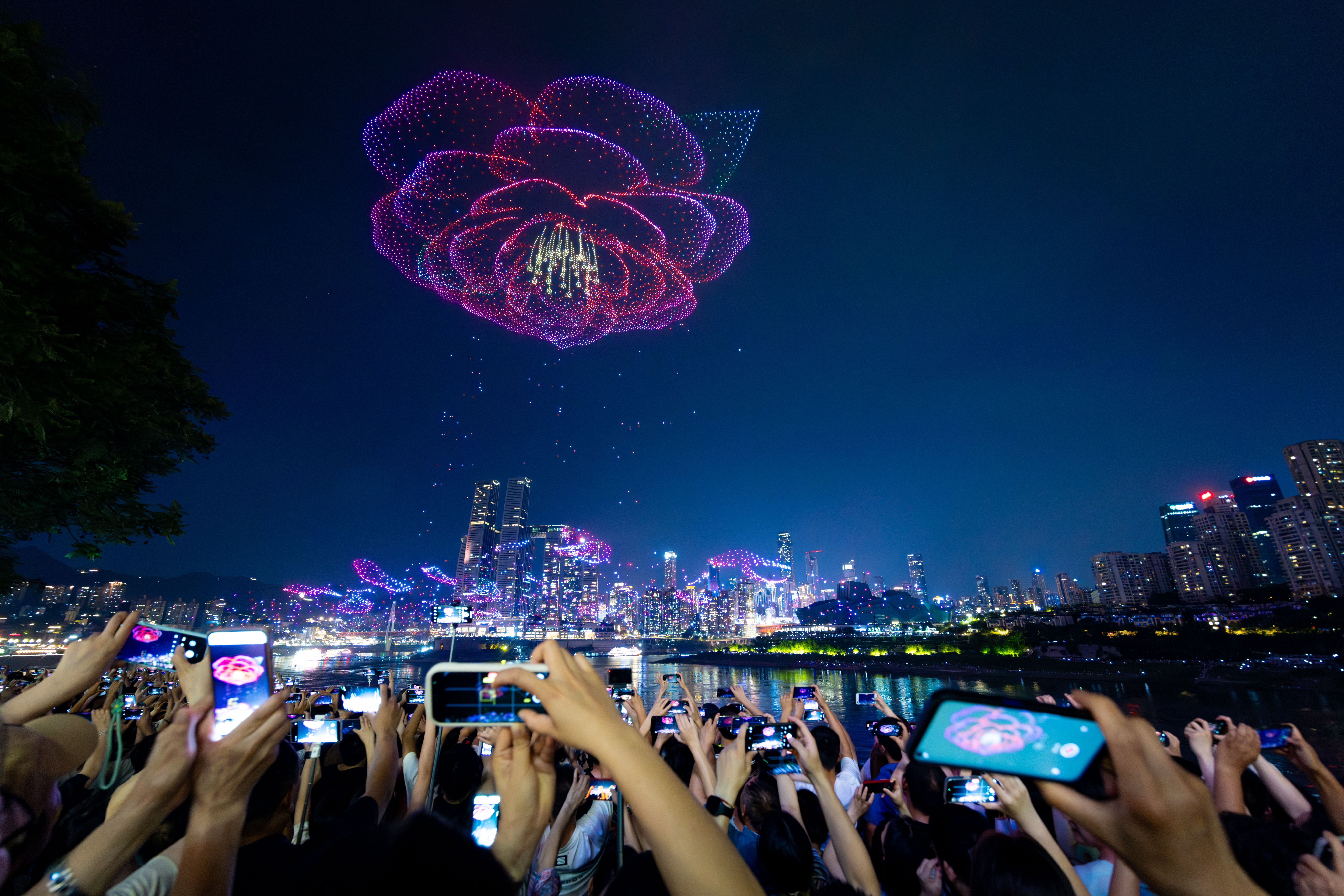 Spectators point their phones toward the sky, taking video and photographs of a drone light show above a city at night. The drones have formed the shape of a blooming flower.