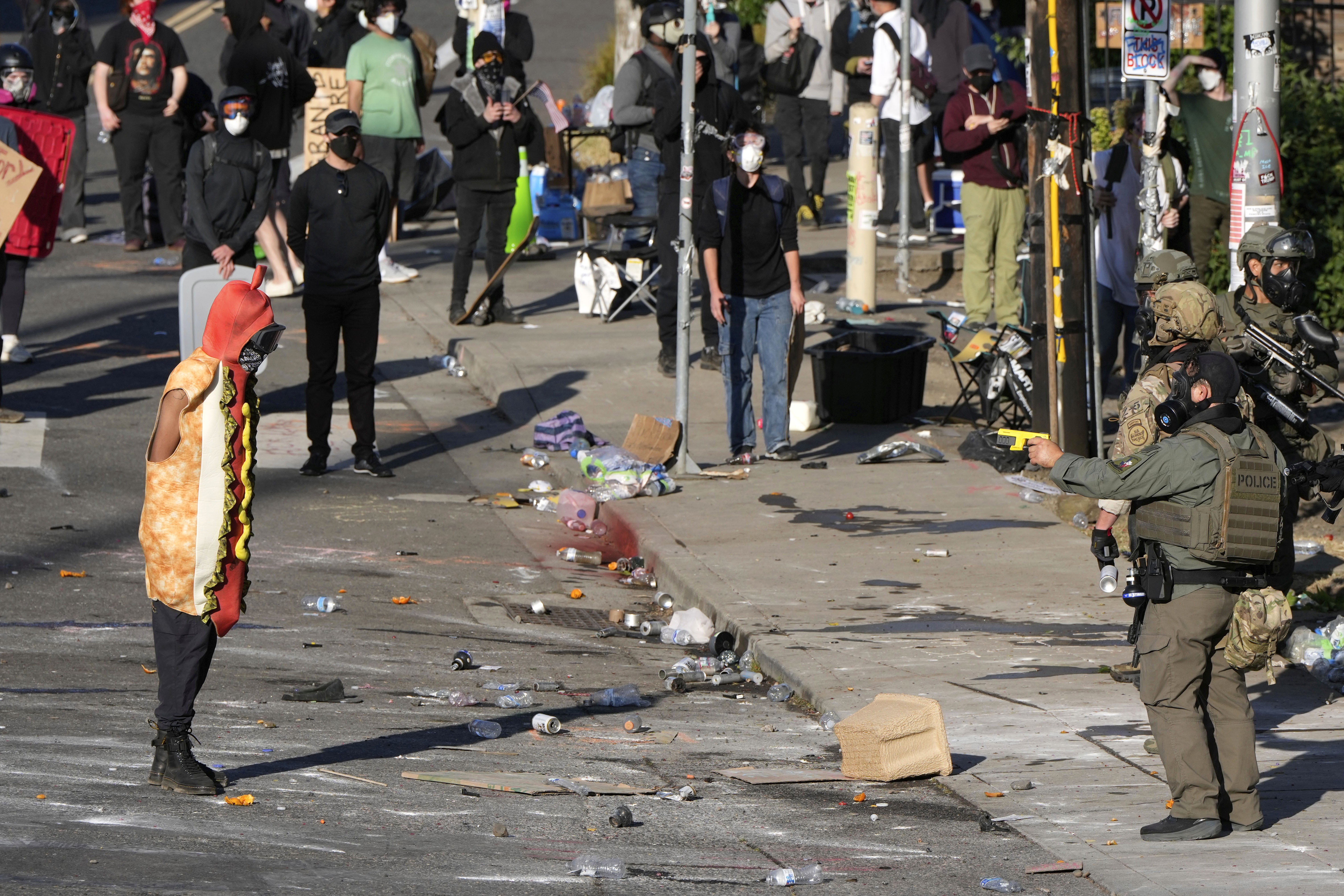 A law enforcement officer points a taser at a person wearing a hot dog costume during a protest.