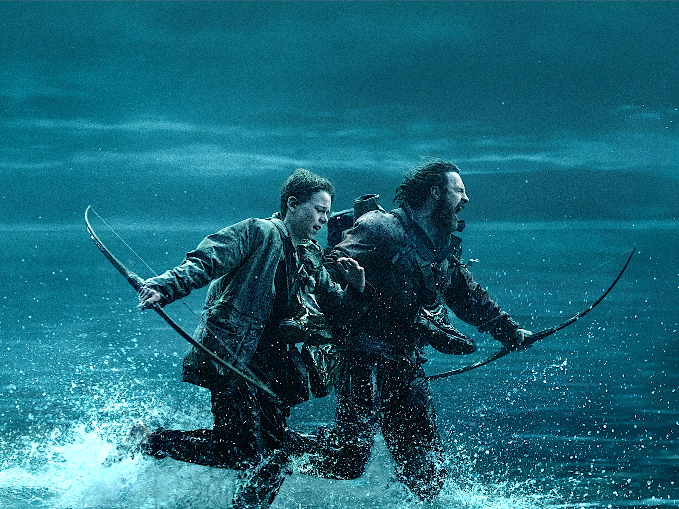 A teenage boy and a man run through the sea at night. They're both dressed in dark clothing and have their boots tied around their necks. They're both holding wooden bows.