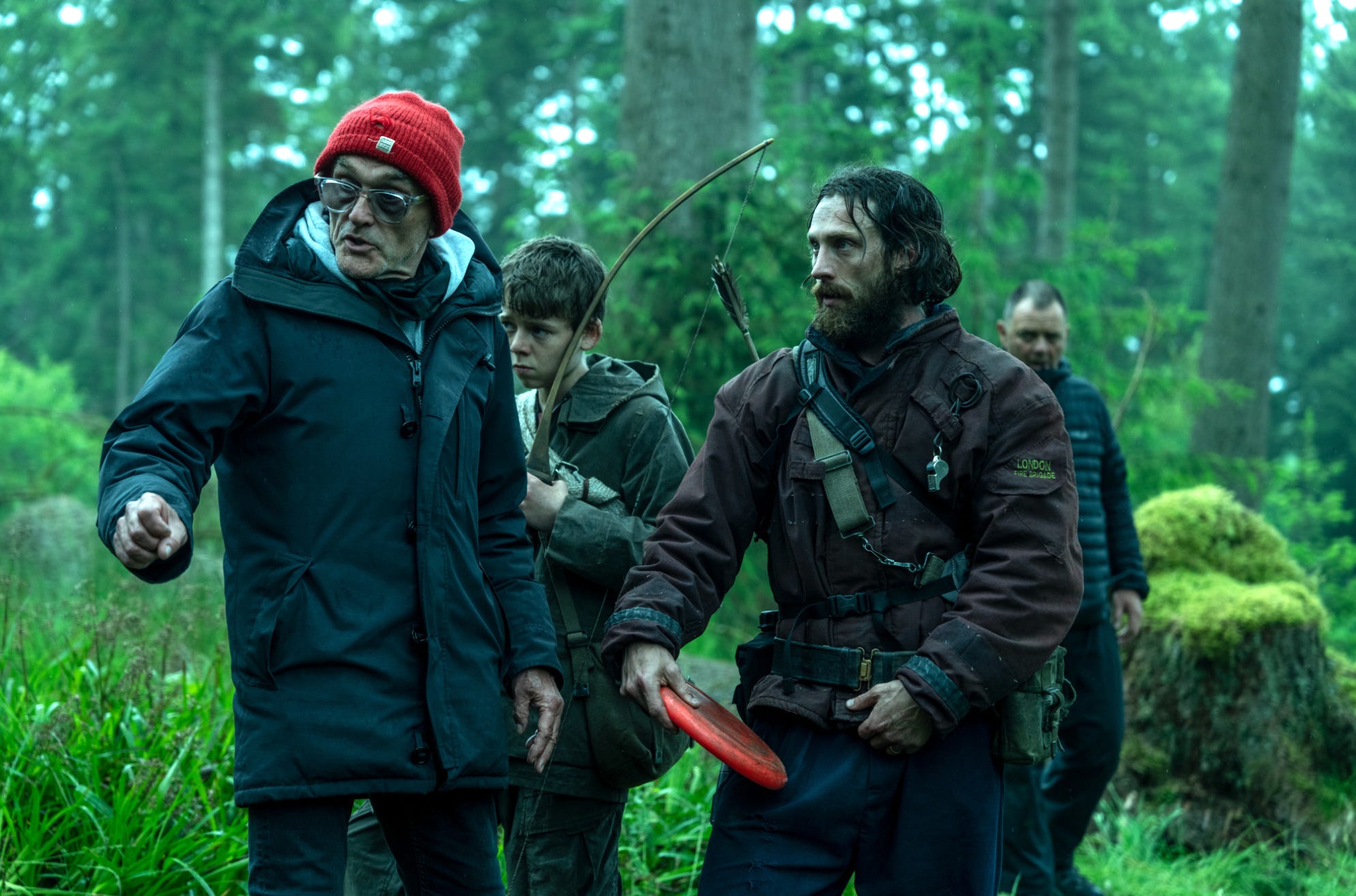 A man in a red bobble hat and a thick black coat is speaking and looks to the left. Behind him is a young boy in a khaki jacket who is holding a wooden bow. Next to him is a man with long wet brown hair and a brown beard. He's wearing a thick red jacket with an arrow hanging over his shoulder. They're stood in a forest.