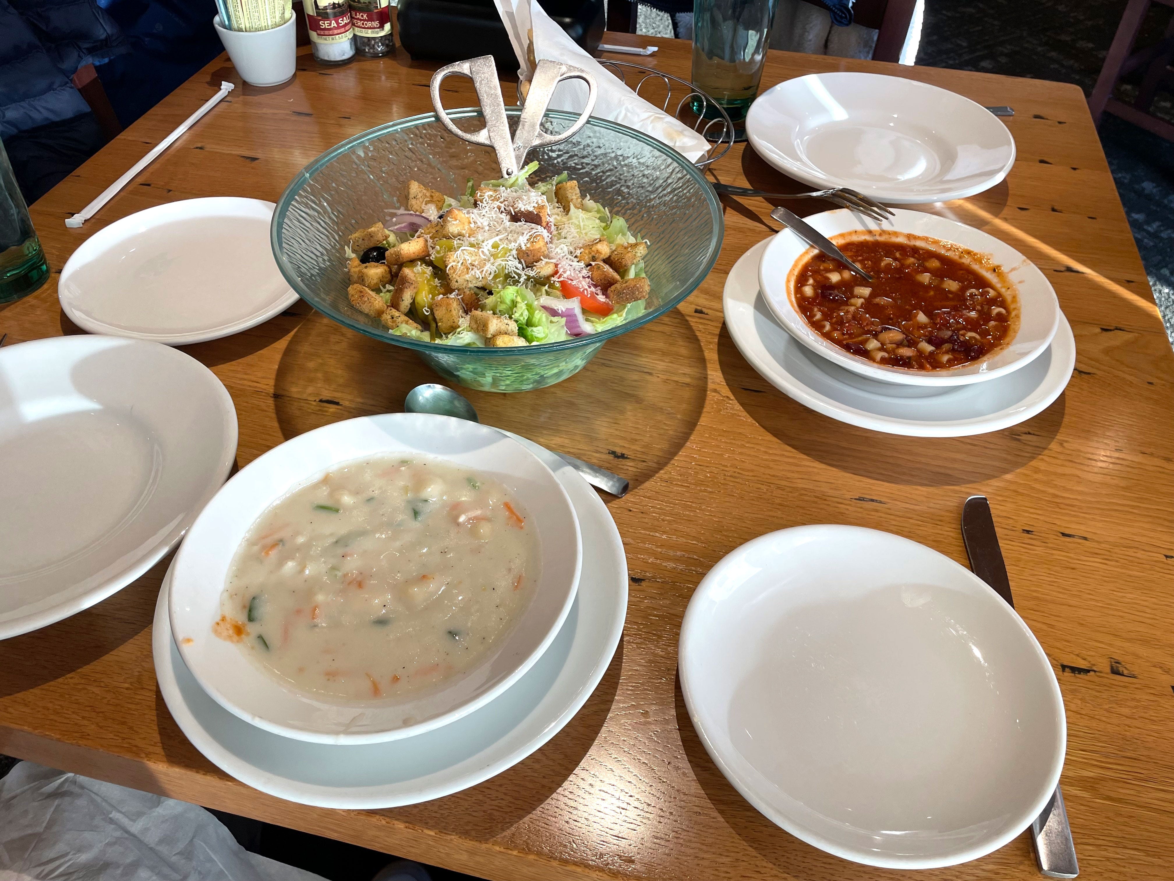 A large bowl of salad, two different soups, and several plates on a table at Olive Garden.