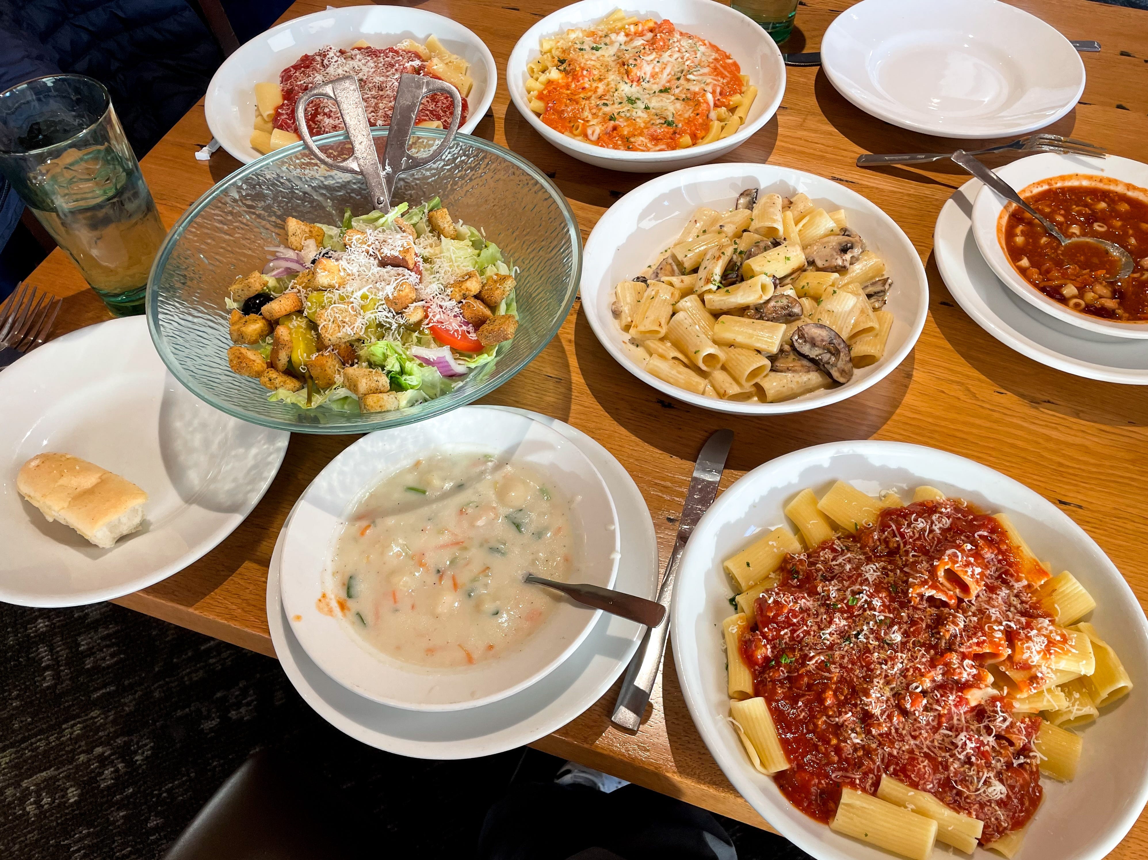 Four different pasta dishes, two different soups, a large bowl of salad, and a half-eaten breadstick on a table at Olive Garden.
