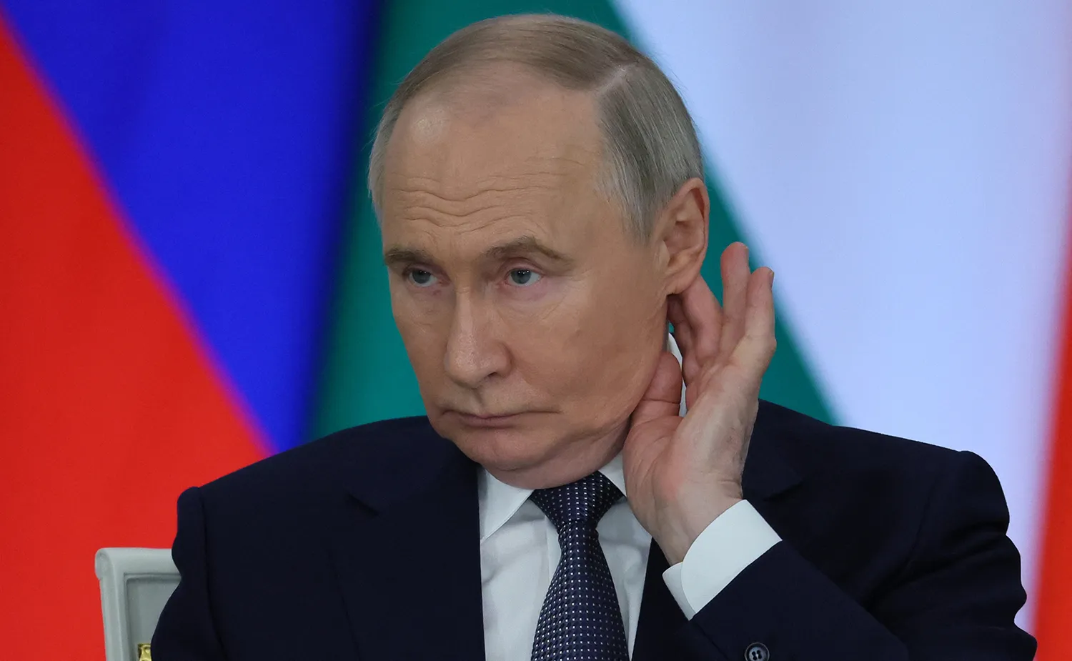 Vladimir Putin puts his hand to his ear as if listening in front of multi colored flags.