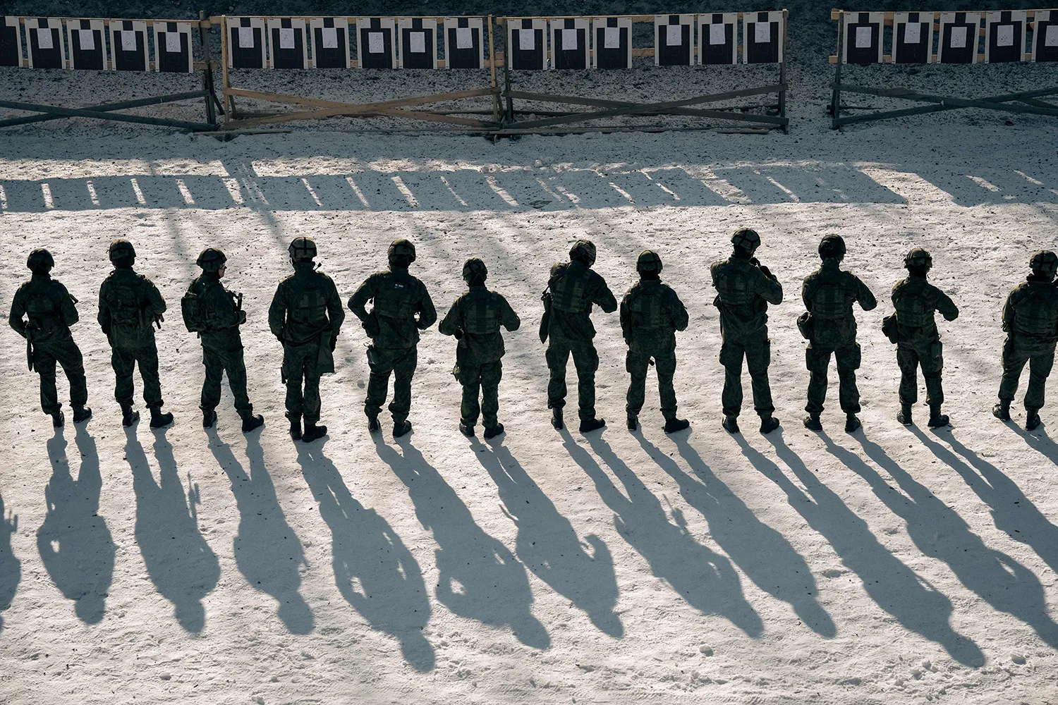 A line of soldiers cast long shadows as they face away toward targets on a shooting range.