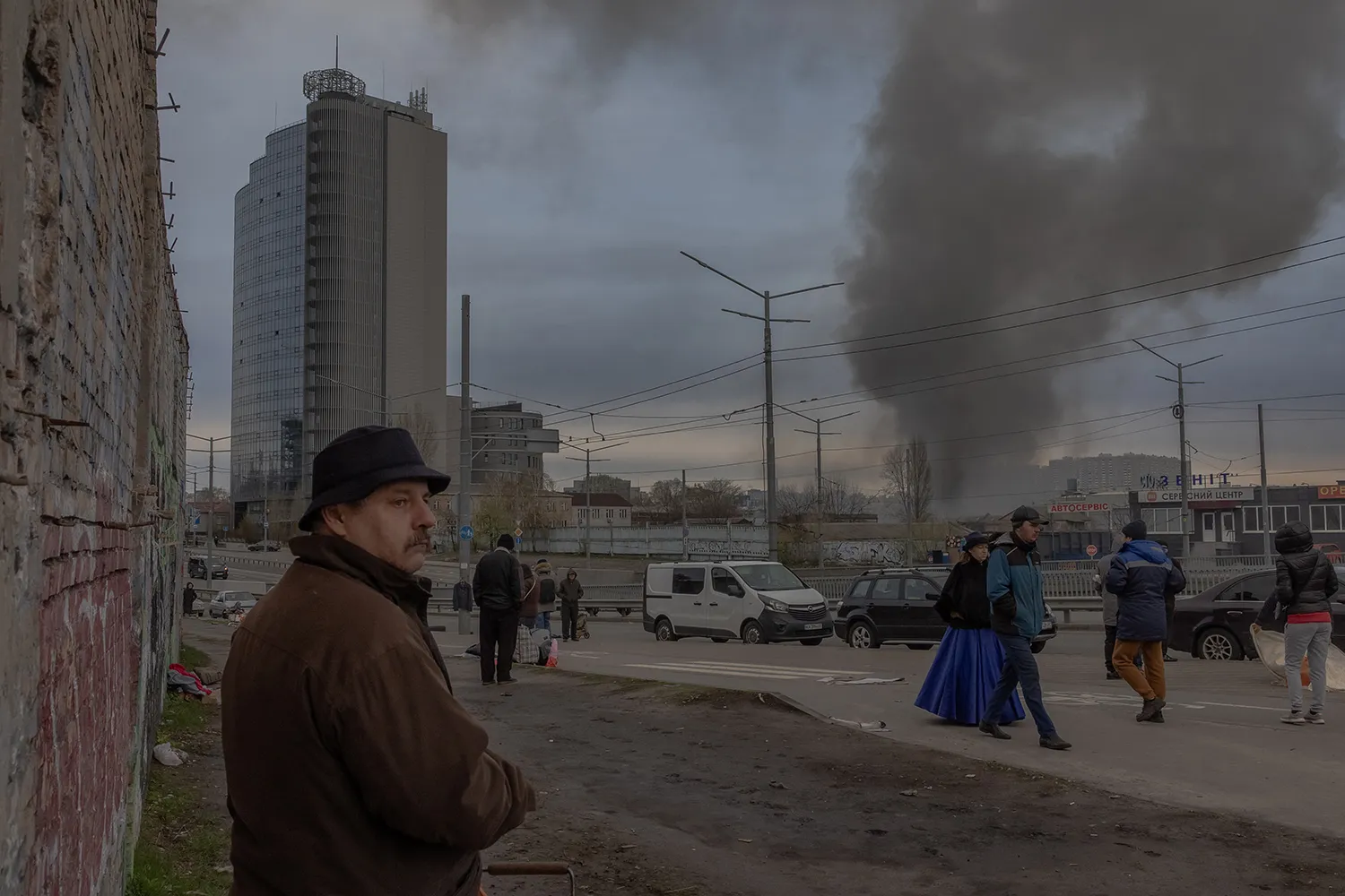Smoke rises in the distance. A tall building is seen at left. Two people walk along a street in the middle distance and a man in the foreground wearing a hat and coat looks up.