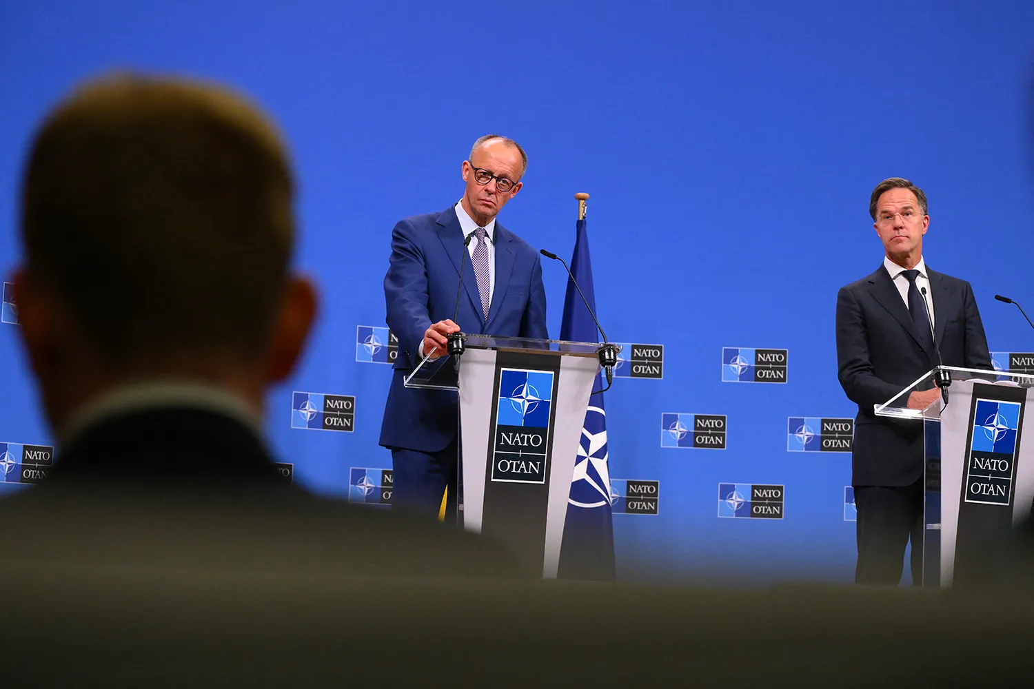 Two men stand at podiums with NATO logos. A blue wall with NATO logos is behind them along with a NATO flag on a stand. An out of focus person back to camera is seen in the foreground.