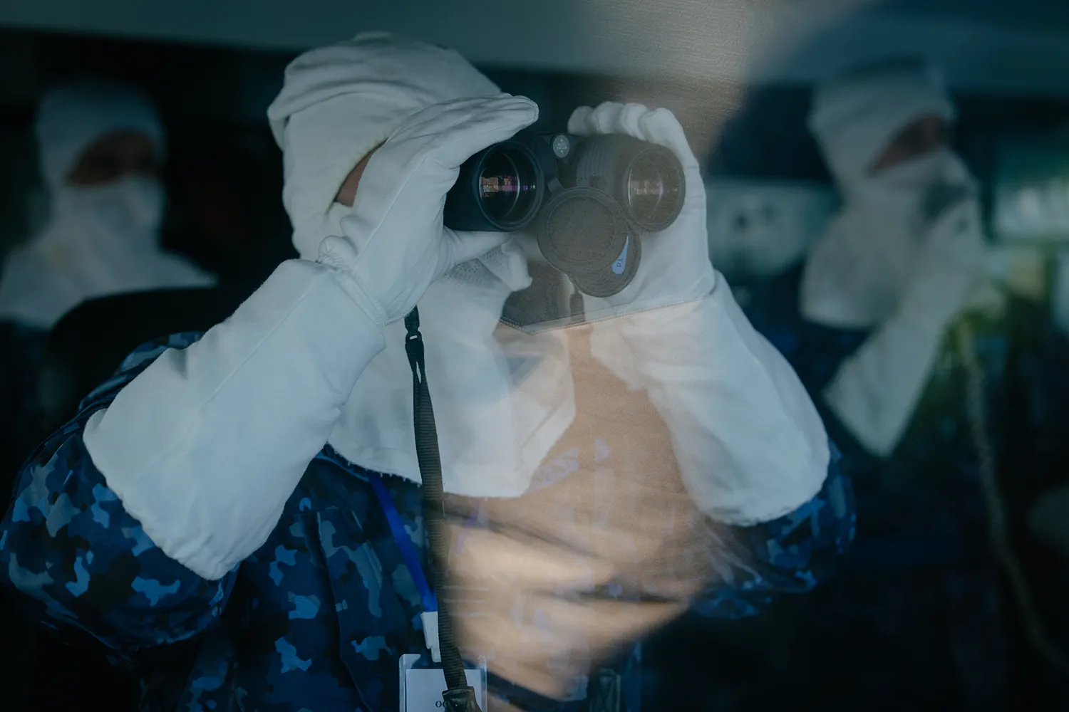A soldier in long white gloves and a white head covering looks through binoculars.