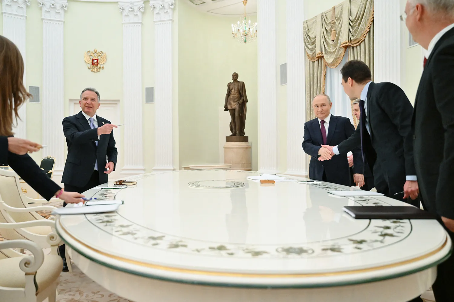 Men gather around a large round table. Witkoff points with one hand across the table toward Putin who is shaking hands with another man.