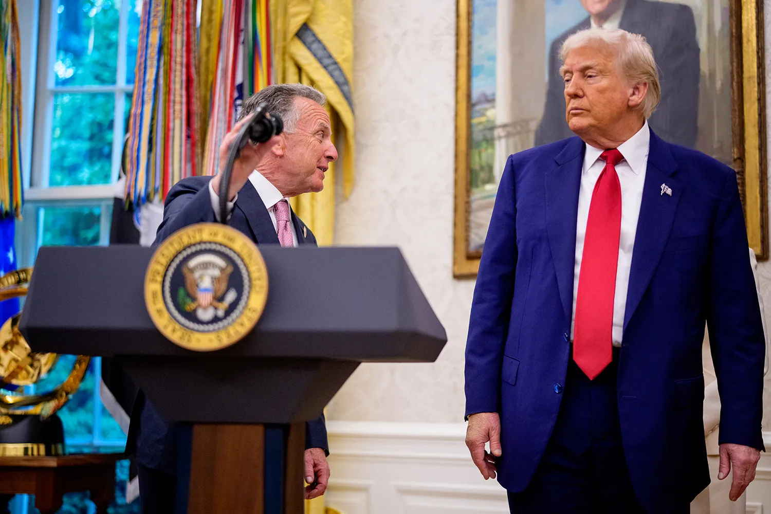 Witkoff adjusts a microphone on a lectern as he turns toward Trump who appears to frown at him.