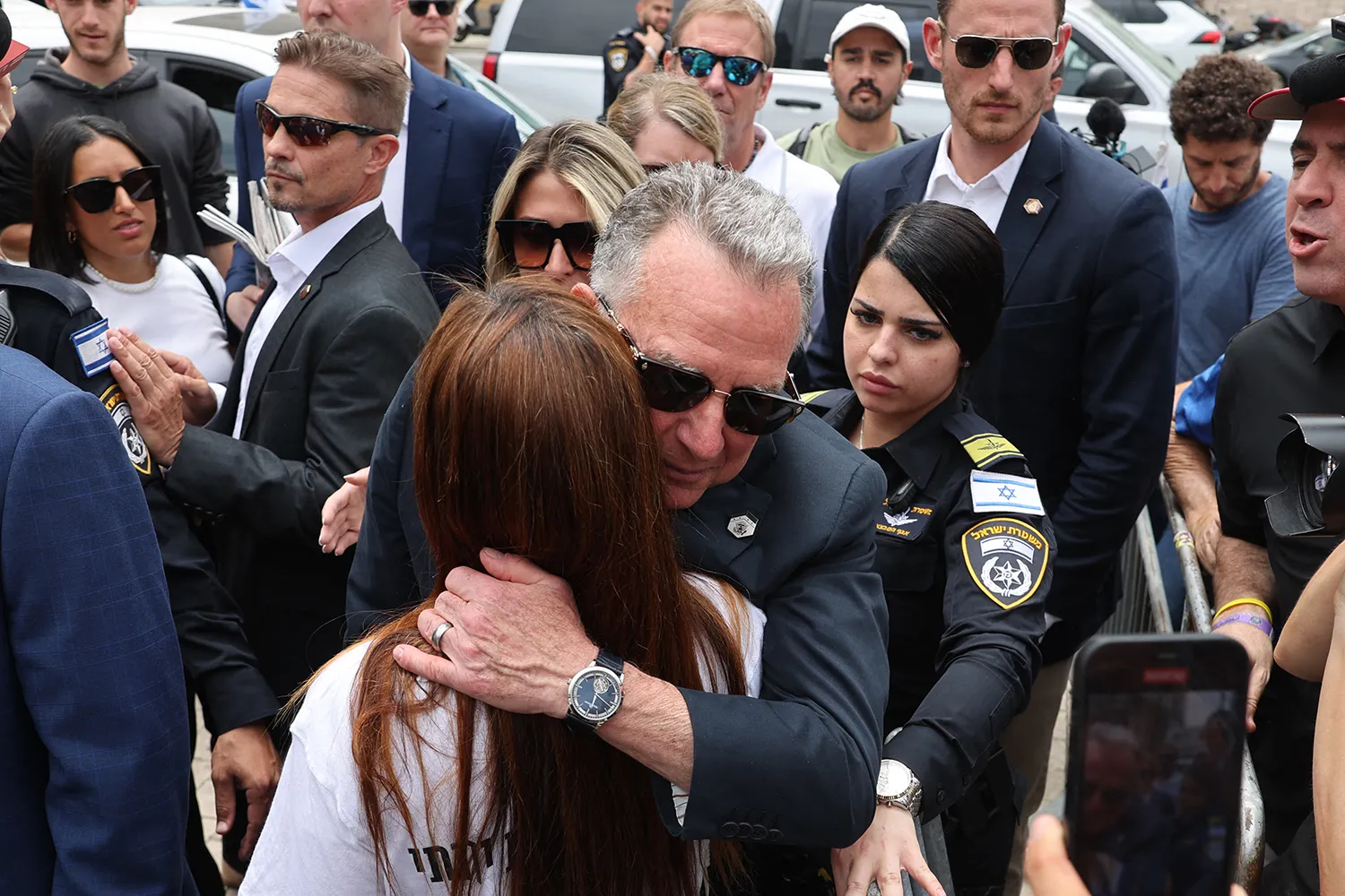 Witkoff wearing sunglasses hugs a woman with long hair as other people gather around them.