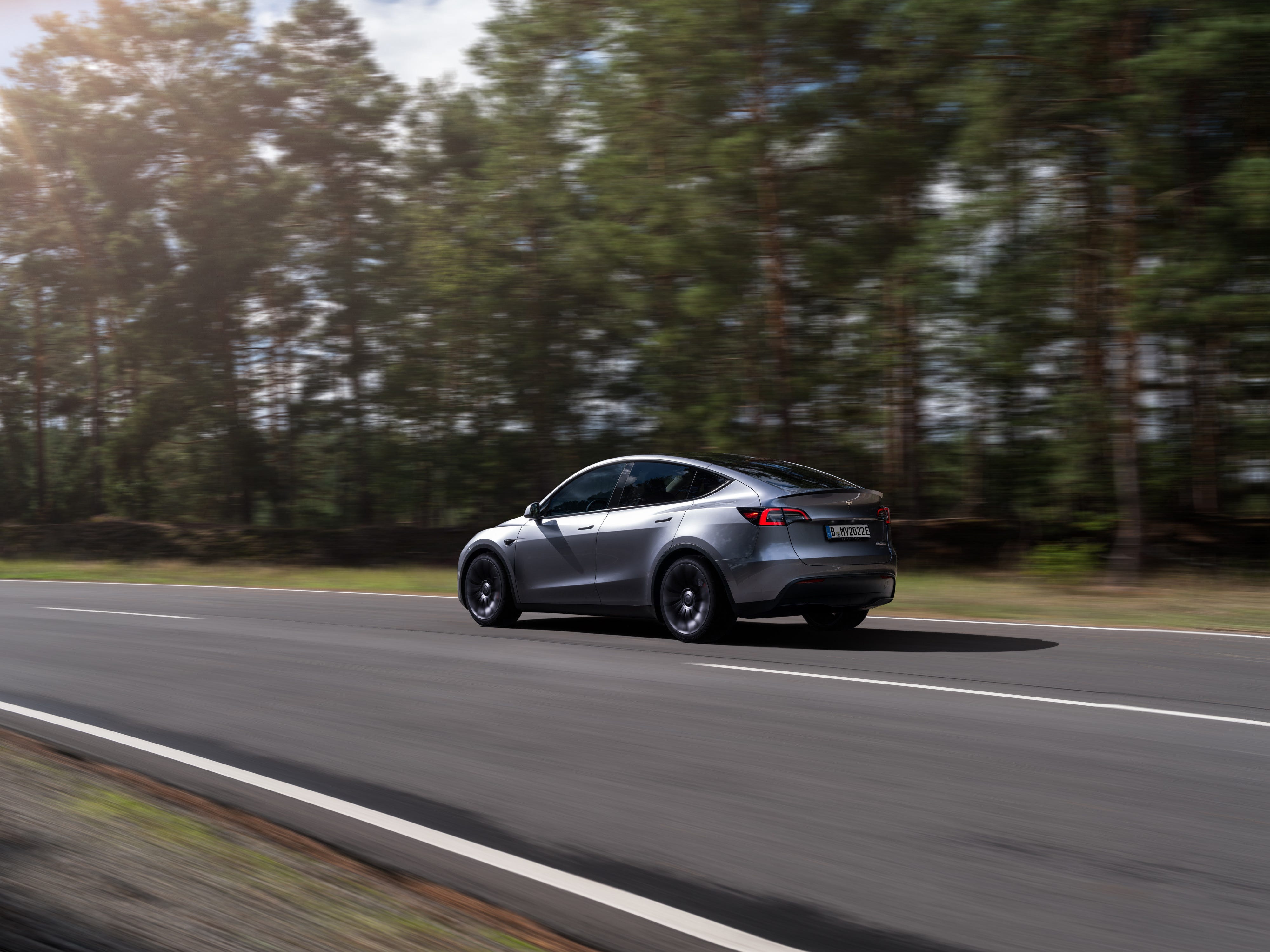 A gray Tesla Model Y SUV is shown driving down the highway from a rear view with trees in the background.