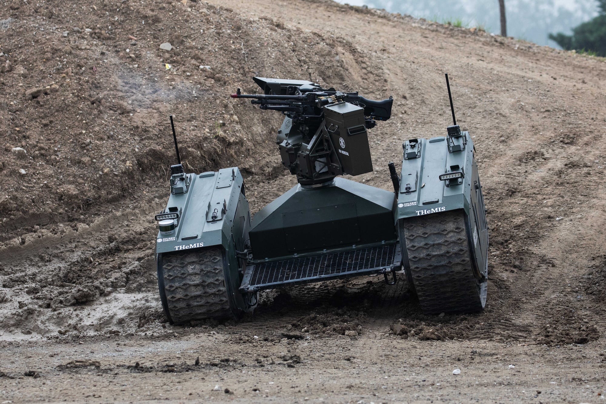 A dark green robot with tracks and a gun on top drives on a muddy hill