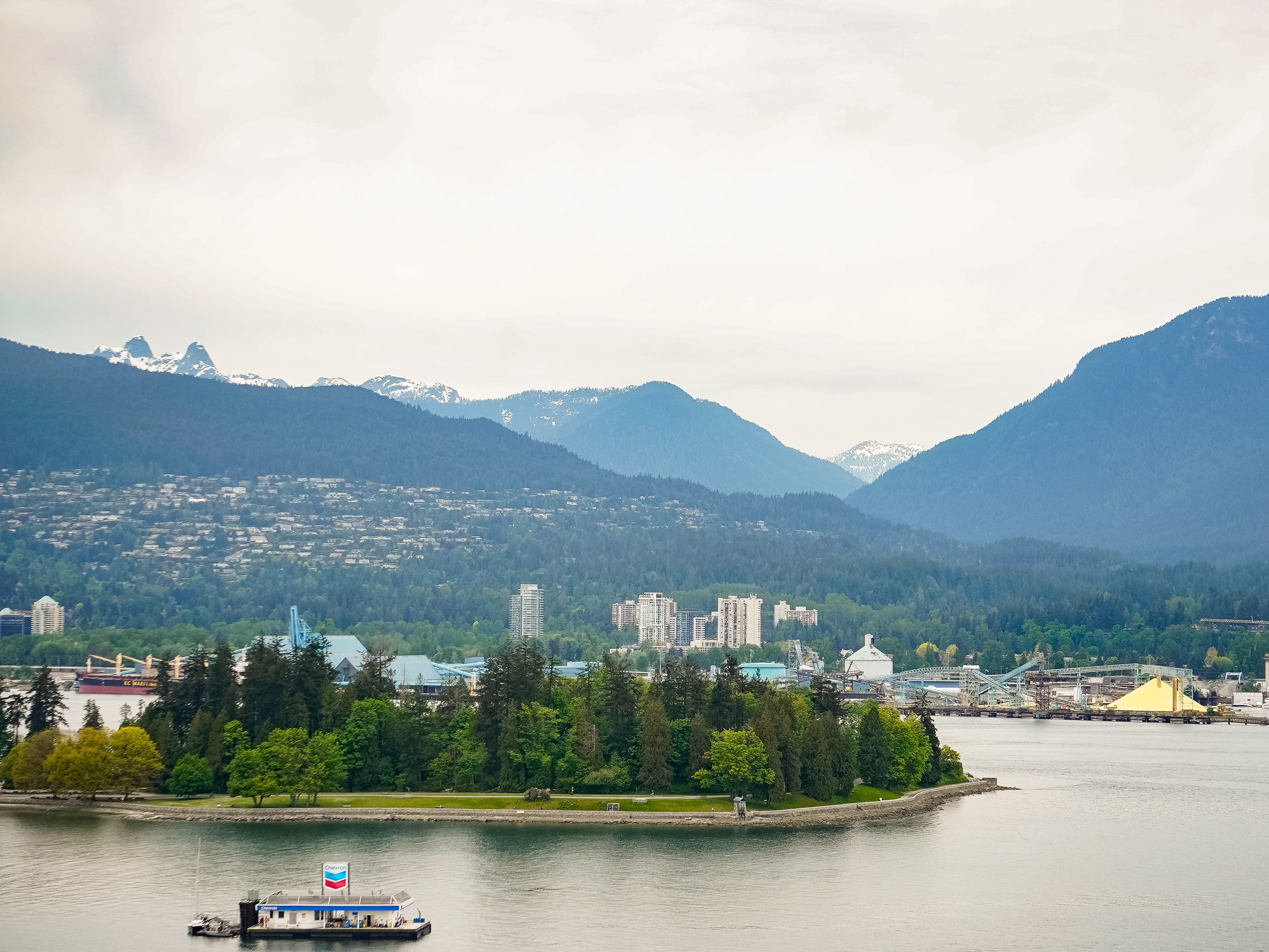 A body of water with a tree-topped island in the middle, a floating gas station in the foreground, and buildings and forests in front of a mountain backdrop